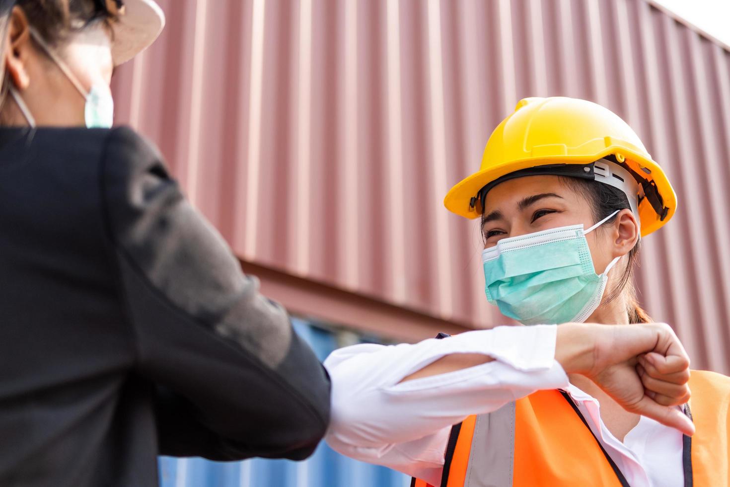 selettivo messa a fuoco a viso di femmina industriale magazzino contenitore lavoratore indossare sicurezza casco e chirurgico viso maschera. saluto con gomito con collega. nuovo normale stile di vita, sociale distanza posto di lavoro foto