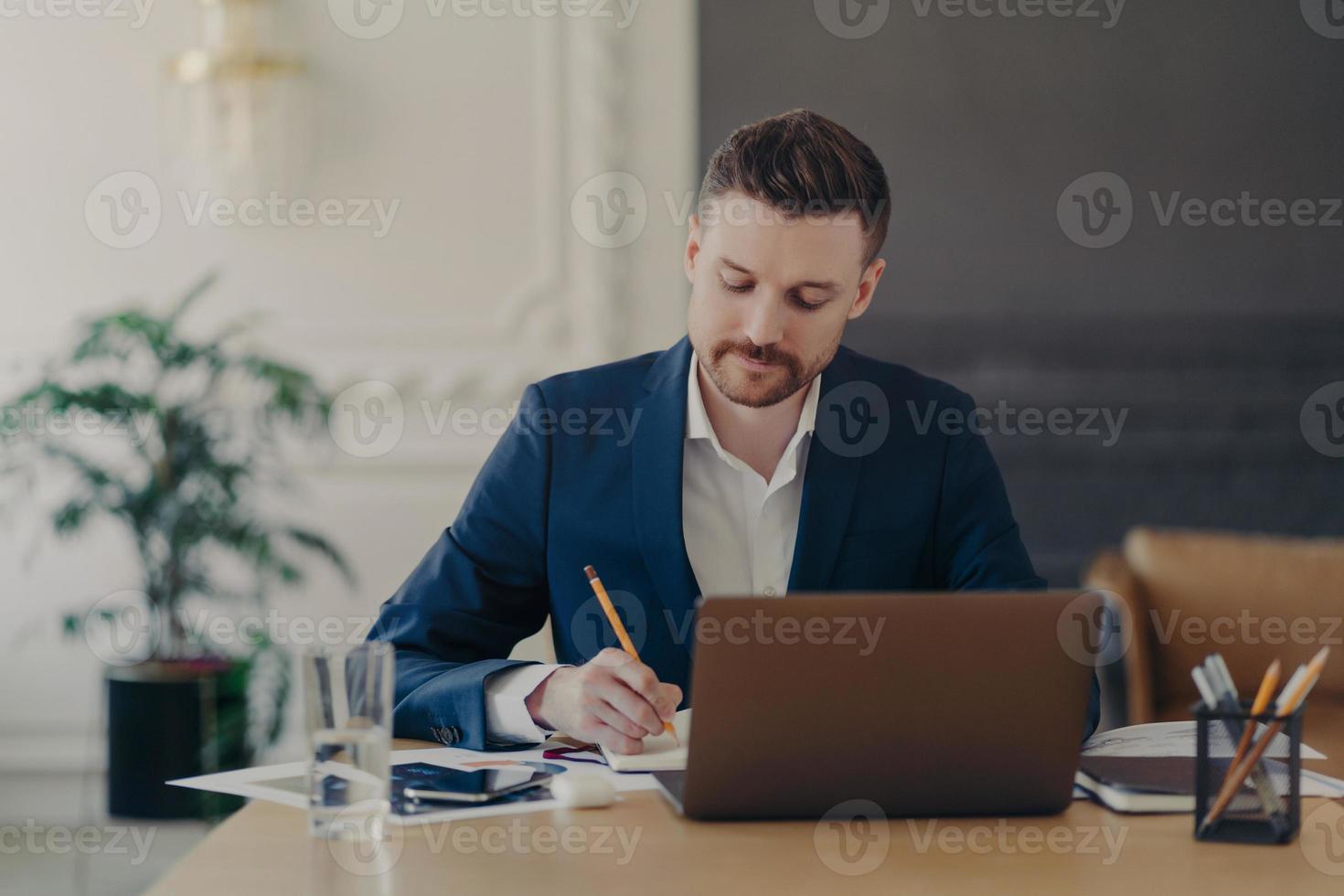 serio concentrato imprenditore maschio esperto controlla il rapporto di contabilità finanziaria scrive alcune note nel blocco note si siede nello spazio di coworking utilizza un computer portatile vestito formalmente. concetto di occupazione foto