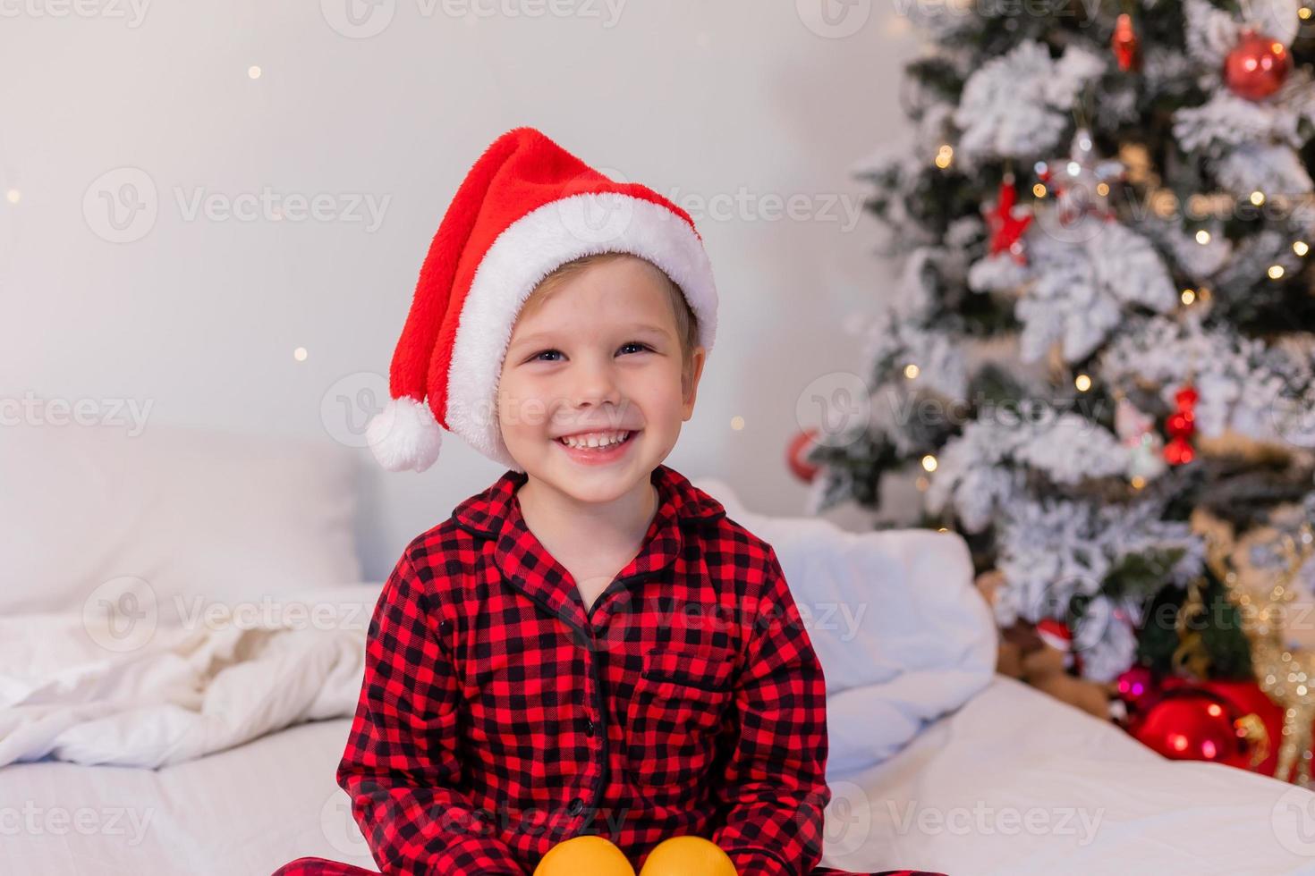 contento poco ragazzo nel pigiama e Santa cappello a casa nel letto mangiare mandarini per Natale foto