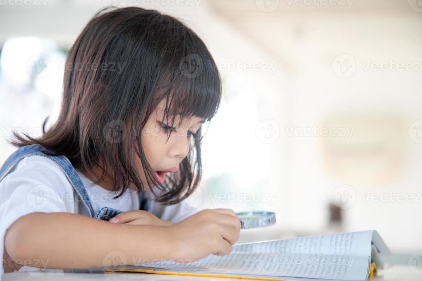 asiatico poco ragazza lettura il libri su il scrivania con un' ingrandimento bicchiere foto