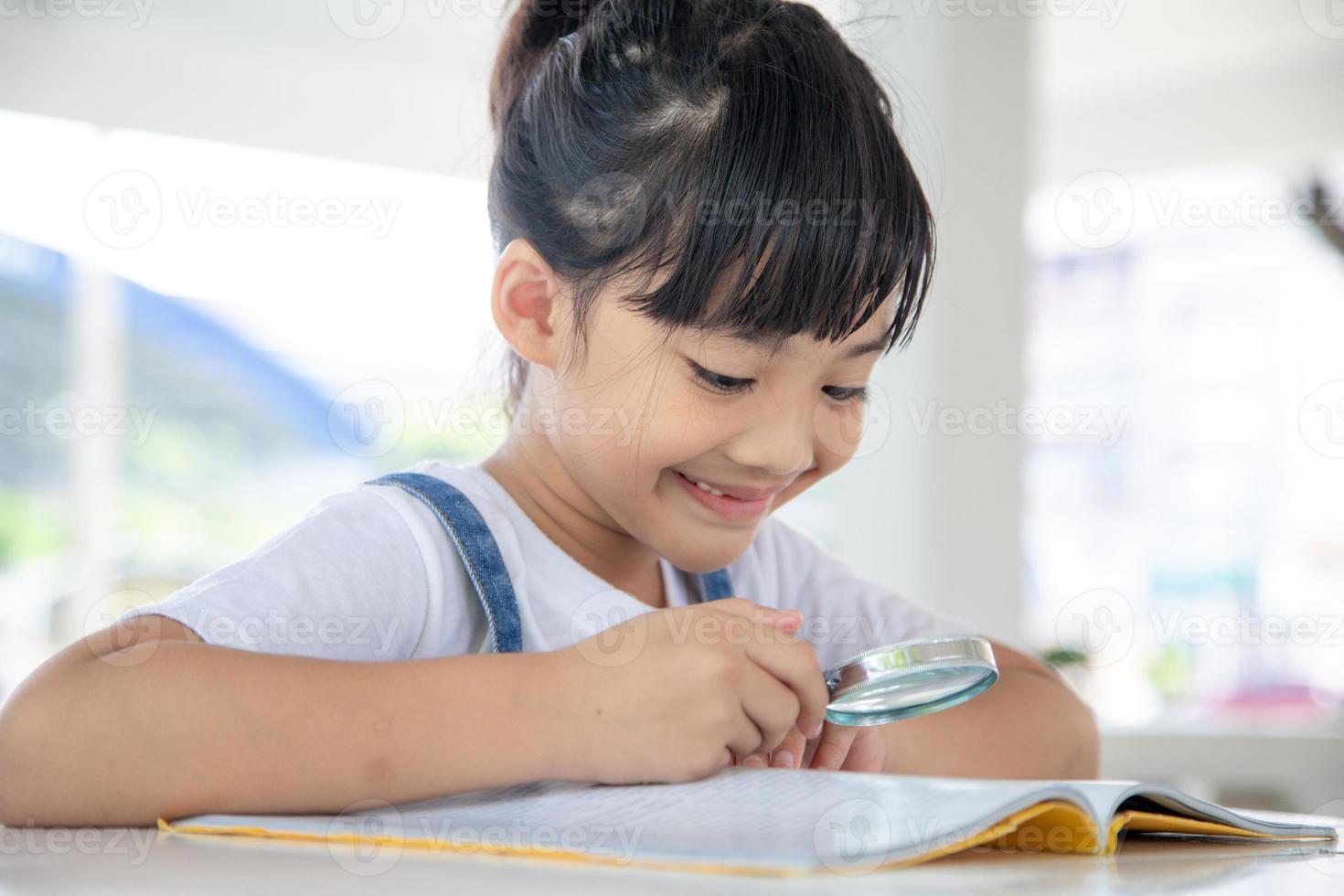 asiatico poco ragazza lettura il libri su il scrivania con un' ingrandimento bicchiere foto