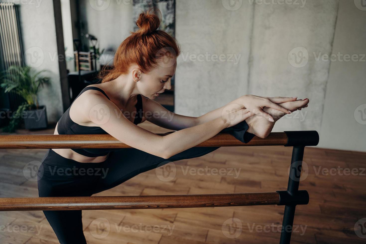 donna dai capelli rossi magra che si allunga sulla sbarra di balletto in classe foto