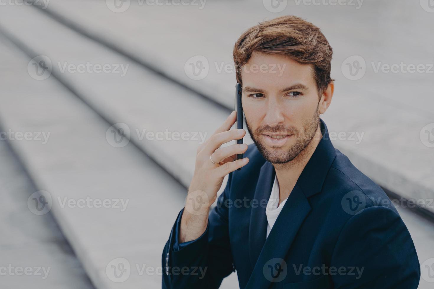 uomo d'affari attraente fiducioso in giacca e cravatta che parla sullo smartphone all'esterno foto
