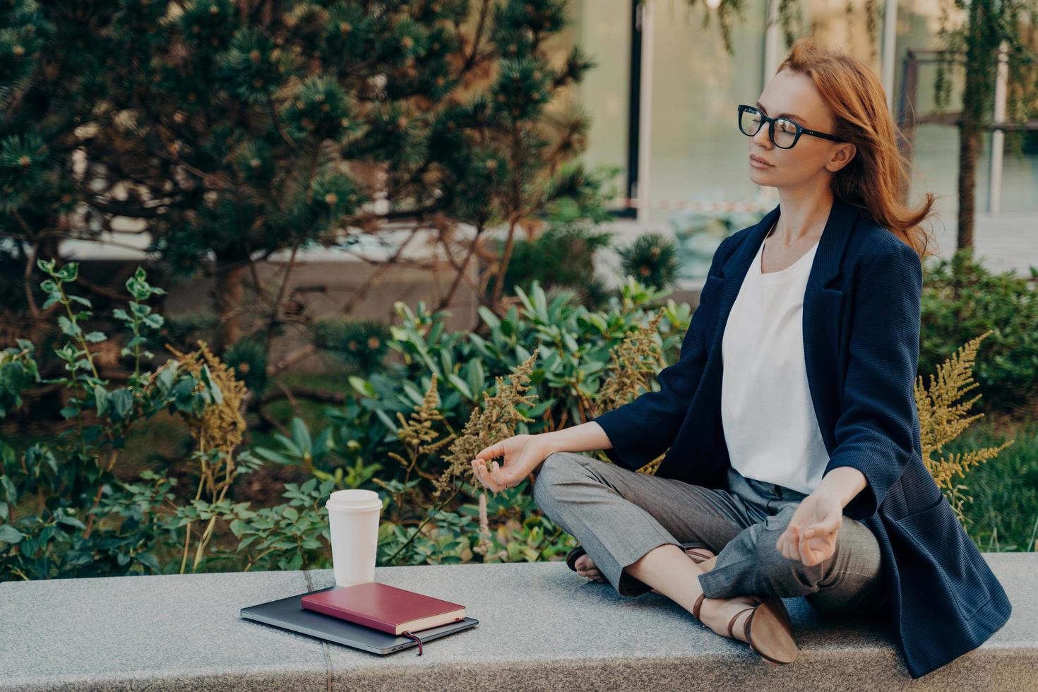 calma donna dai capelli rossi che si rilassa meditando mentre lavora in remoto sul laptop all'aperto foto