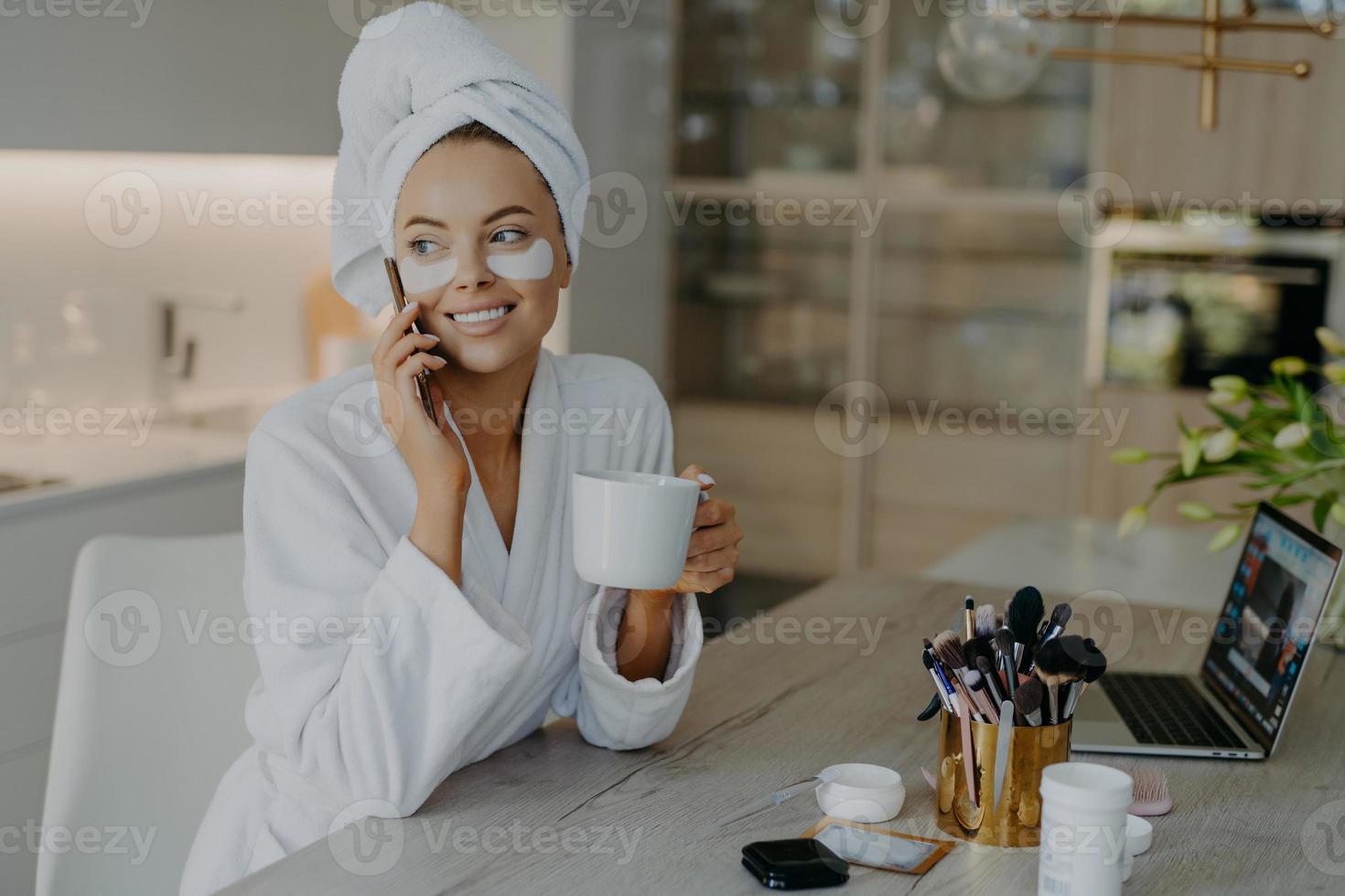 bella donna sorridente applica cerotti idrogel sotto gli occhi si diverte a pulirsi dopo la doccia vestita in accappatoio beve il tè ha una conversazione telefonica si siede sulla sedia a casa. procedure per la cura del viso foto