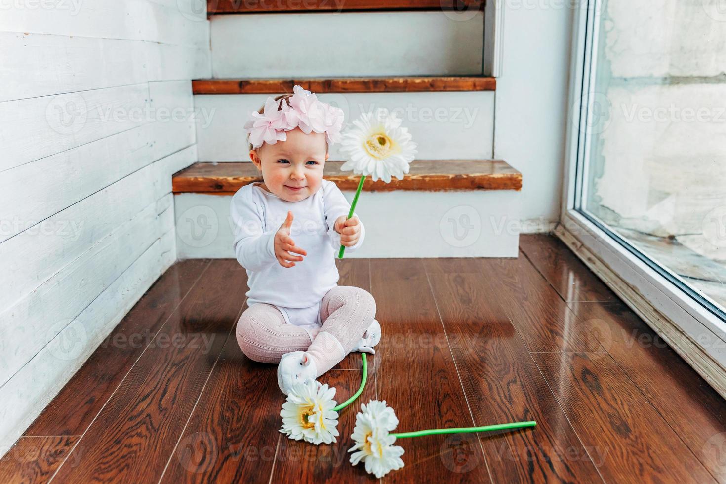 poco sorridente bambino ragazza uno anno vecchio indossare primavera ghirlanda ubicazione su pavimento nel luminosa leggero vivente camera vicino finestra e giocando con gerbera fiori. contento ragazzo giocando a casa. infanzia concetto. foto