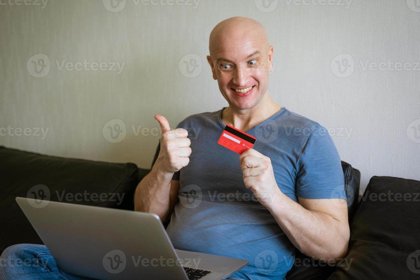 emotivo Calvo uomo su divano con il computer portatile e credito carta nel mano foto