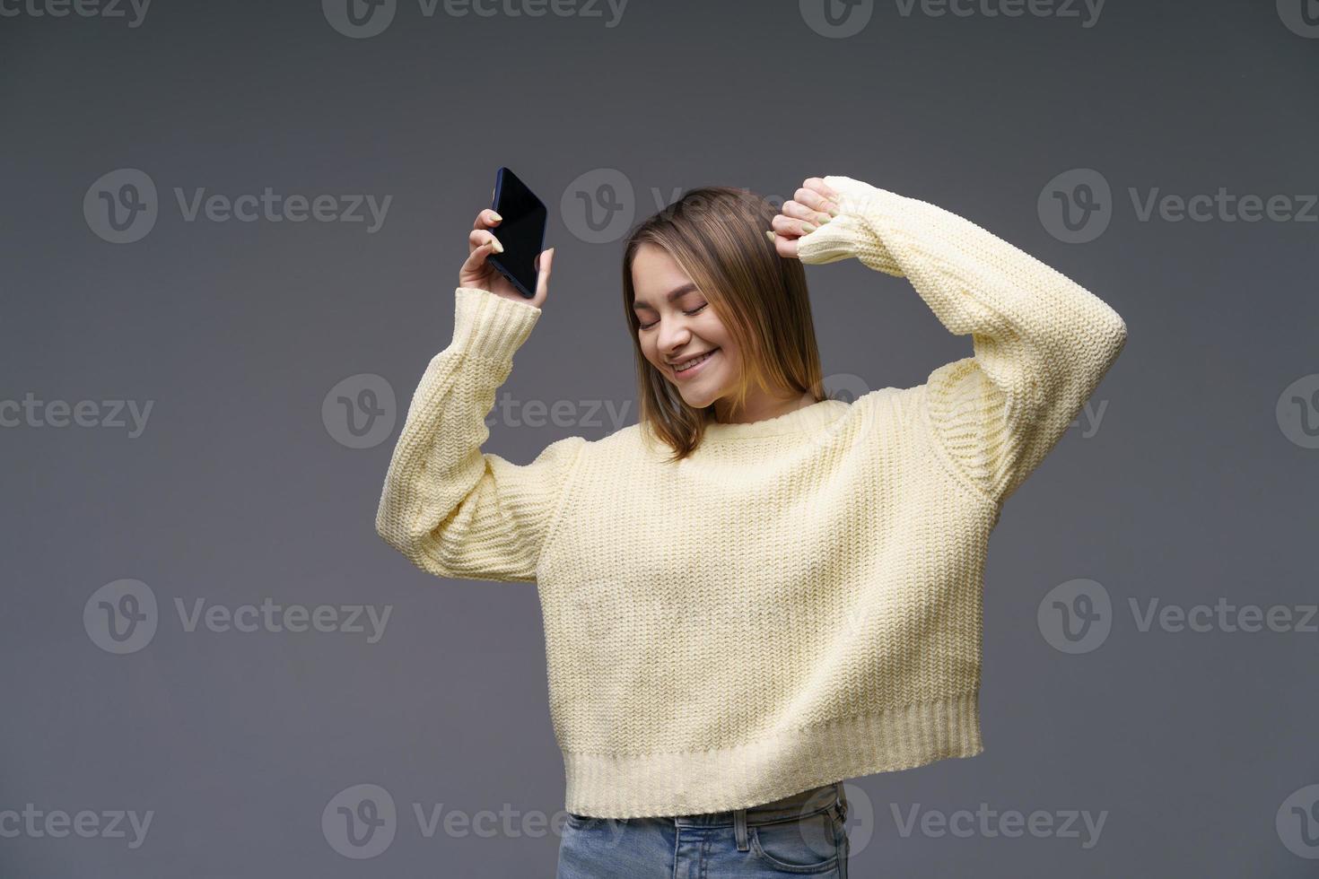 giovane donna nel giallo maglione su grigio sfondo danza con Telefono nel sua mano foto