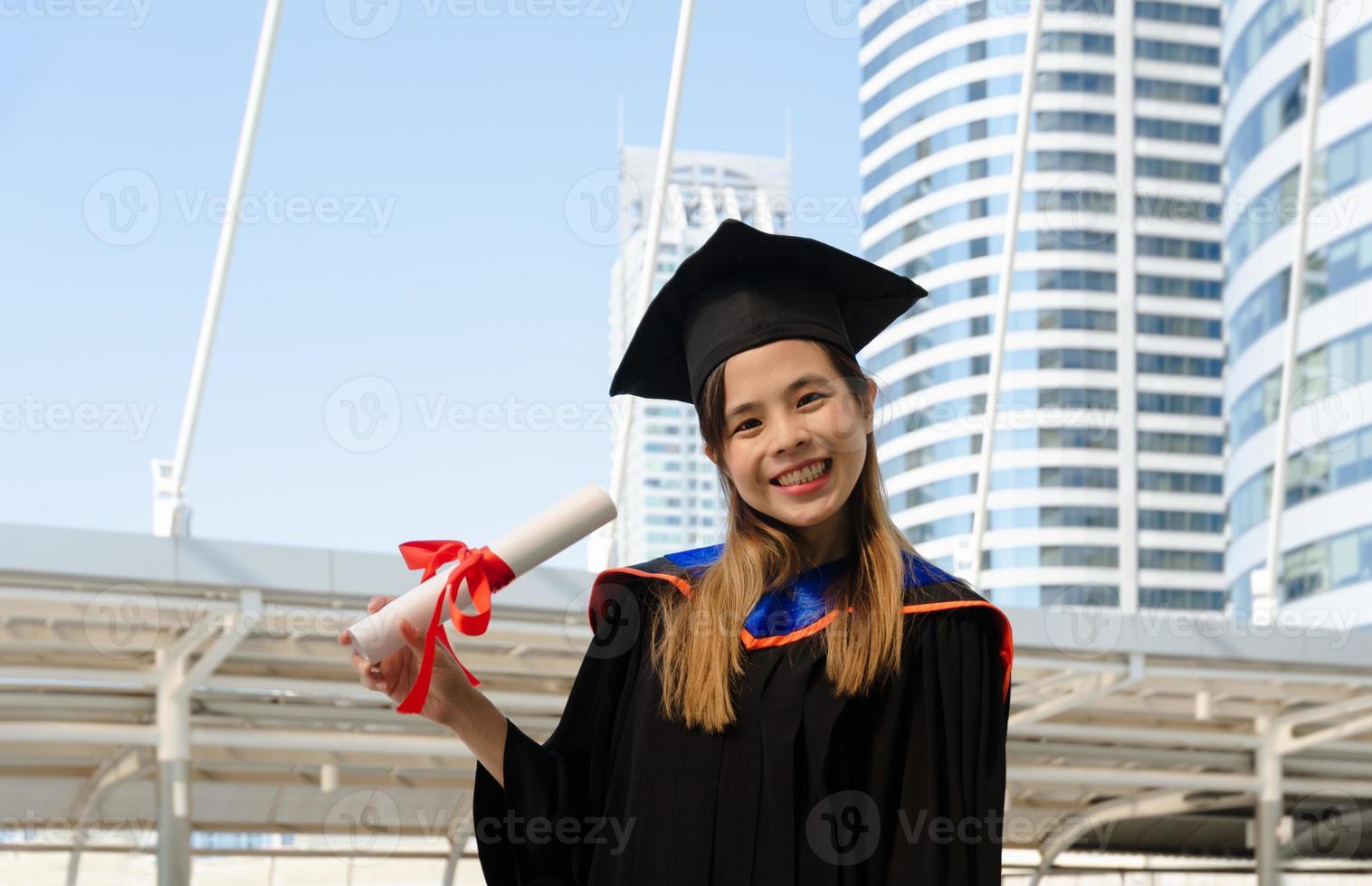 sorridente femmina nel la laurea toga Tenere accademico grado e guardare a telecamera con edificio sfondo foto