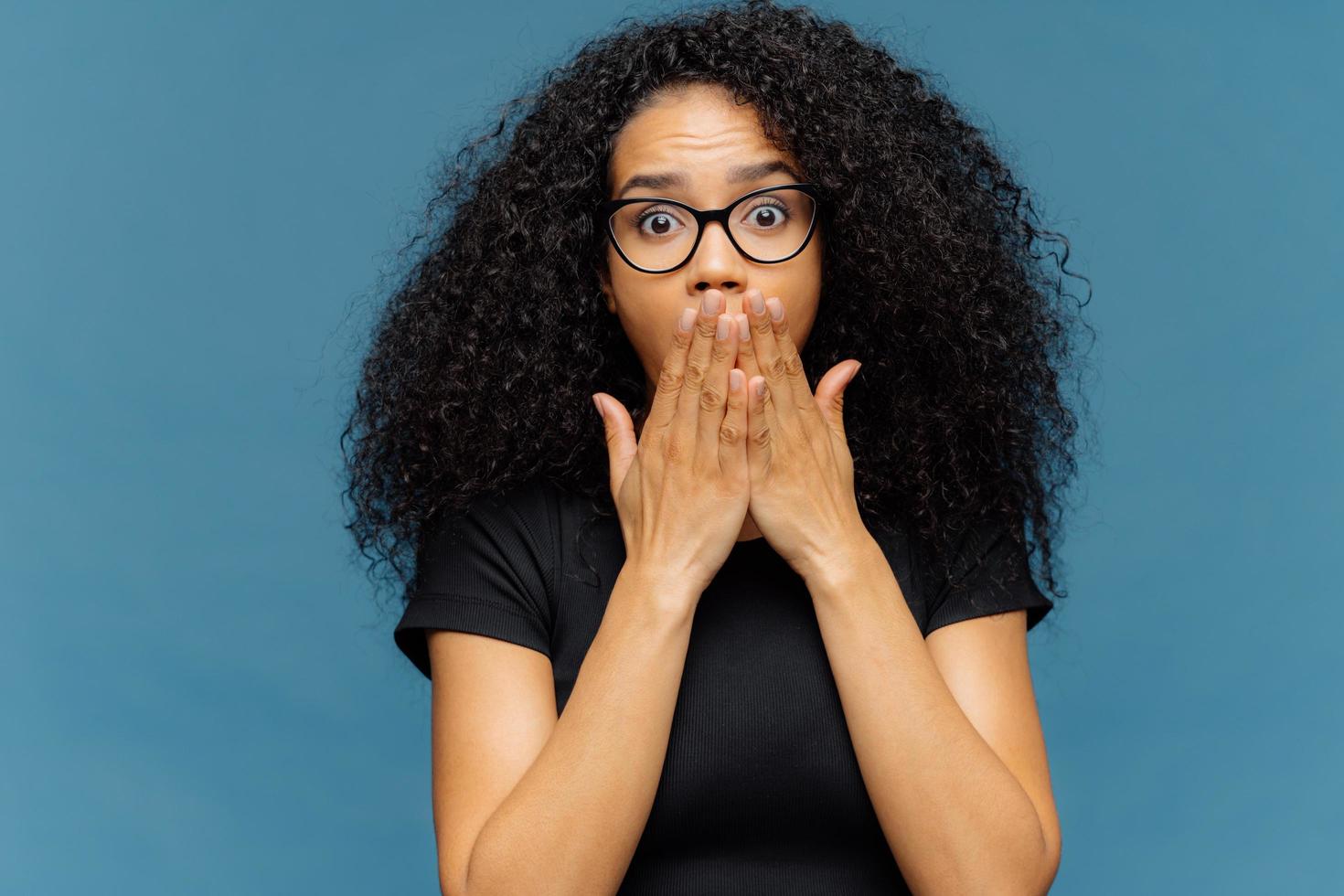 la foto di una giovane donna stupefatta copre la bocca, ha il respiro affannoso, gli occhi stropicciati, i capelli afro, non riesce a credere a notizie scioccanti, indossa una maglietta nera casual, si trova su sfondo blu. omg concetto.