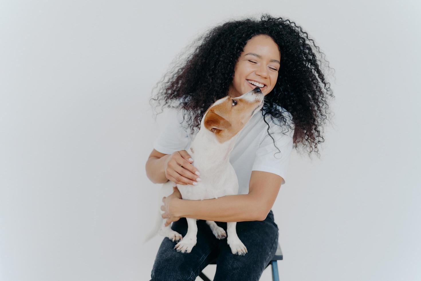 colpo isolato di donna afroamericana sorridente con l'acconciatura riccia, riceve un bacio dal cane preferito, si siede su una sedia contro il muro bianco, indossa maglietta e jeans. persone, animali, emozioni e concetto di amore foto