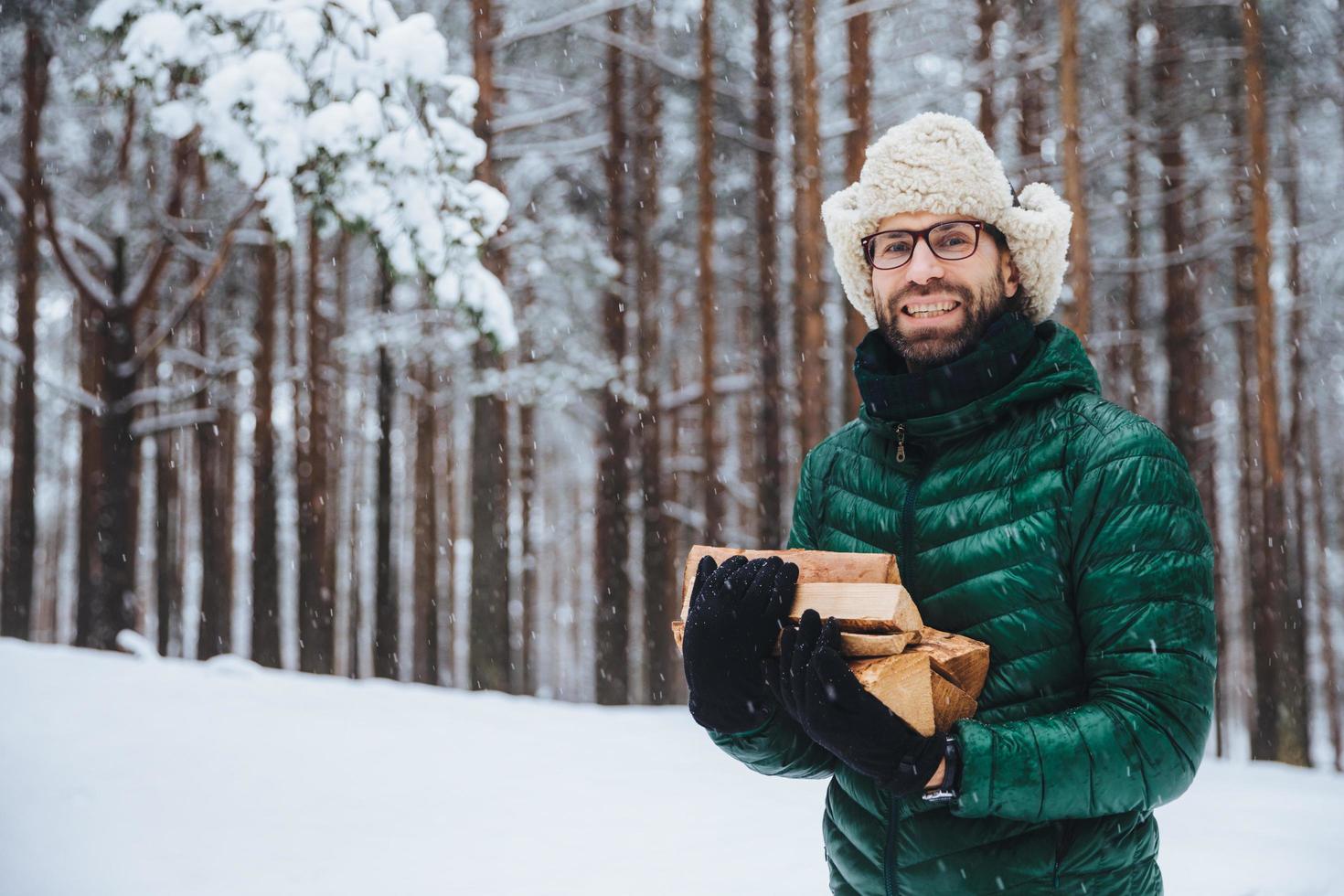 cheerfu barba dall'aspetto gradevole, il modello maschile tiene legna da ardere, trascorre il tempo libero nella foresta, va a friggere shish kebab, fa picnic nella cerchia familiare, taglia legna, gode dell'aria fresca della foresta invernale all'aperto foto