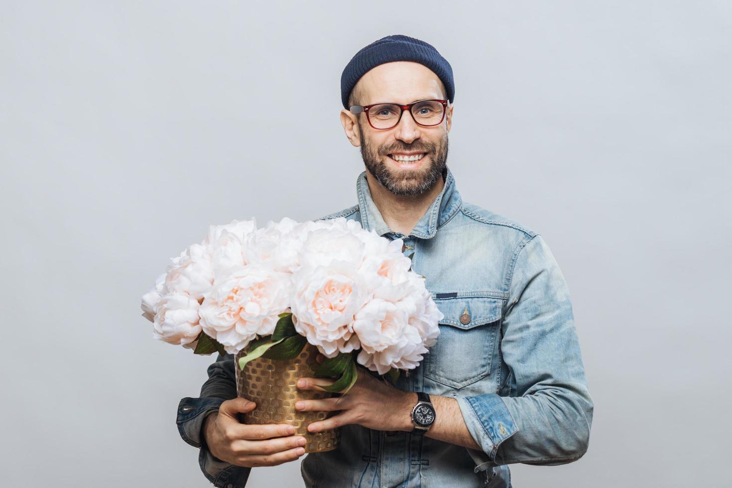 il maschio barbuto soddisfatto con un'espressione allegra ha un ampio sorriso, tiene un mazzo di fiori, indossa un cappello e una giacca di jeans, isolato su sfondo bianco. l'uomo sorridente soddisfatto pone al coperto con i fiori foto