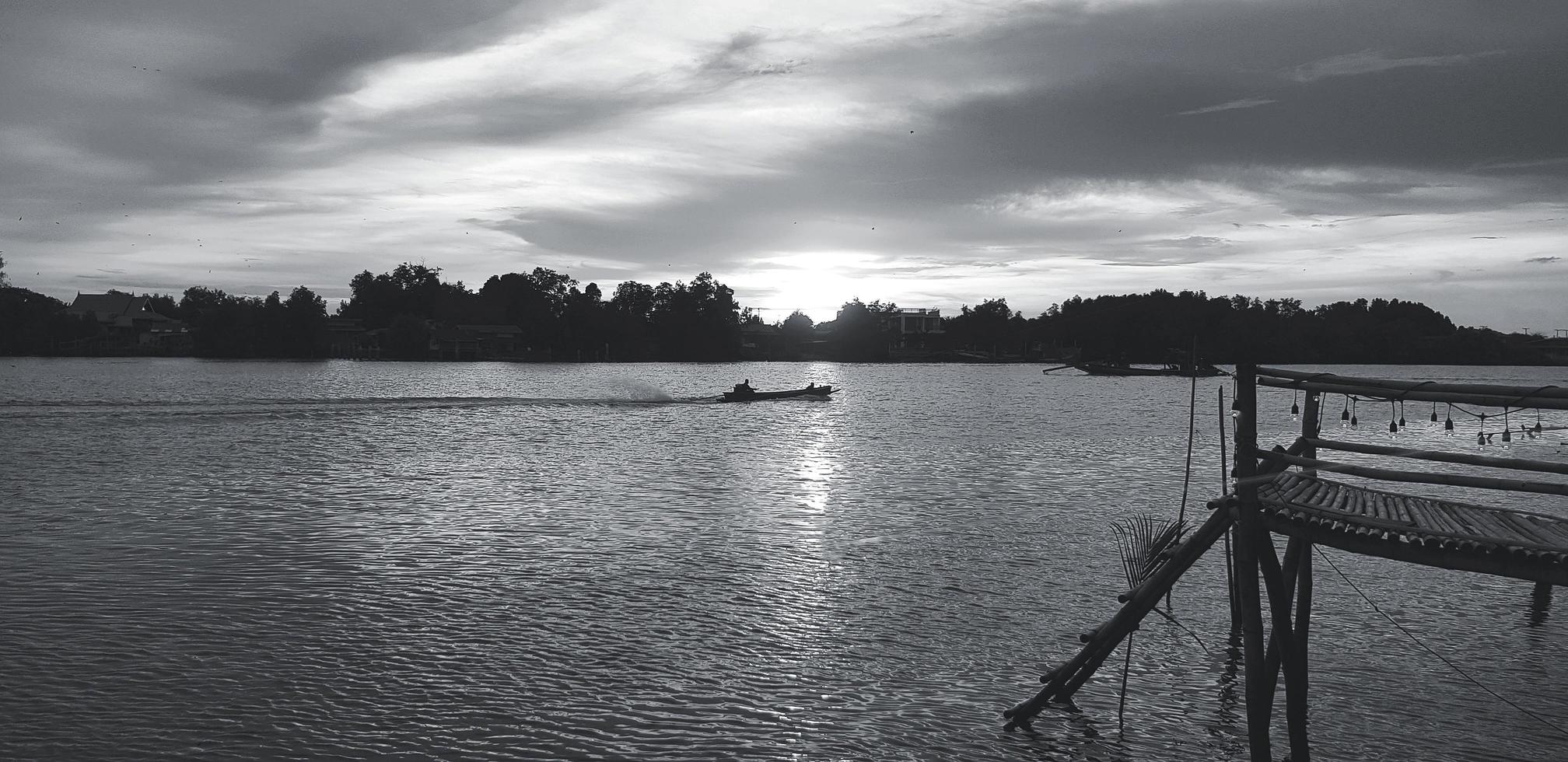 silhouette di di legno riva, lungomare o ponte con coda lunga barca su rive e albero sfondo a tramonto o luce del sole volta. bellissimo natura e trasporto nel nero e bianca tono o monocromo. foto