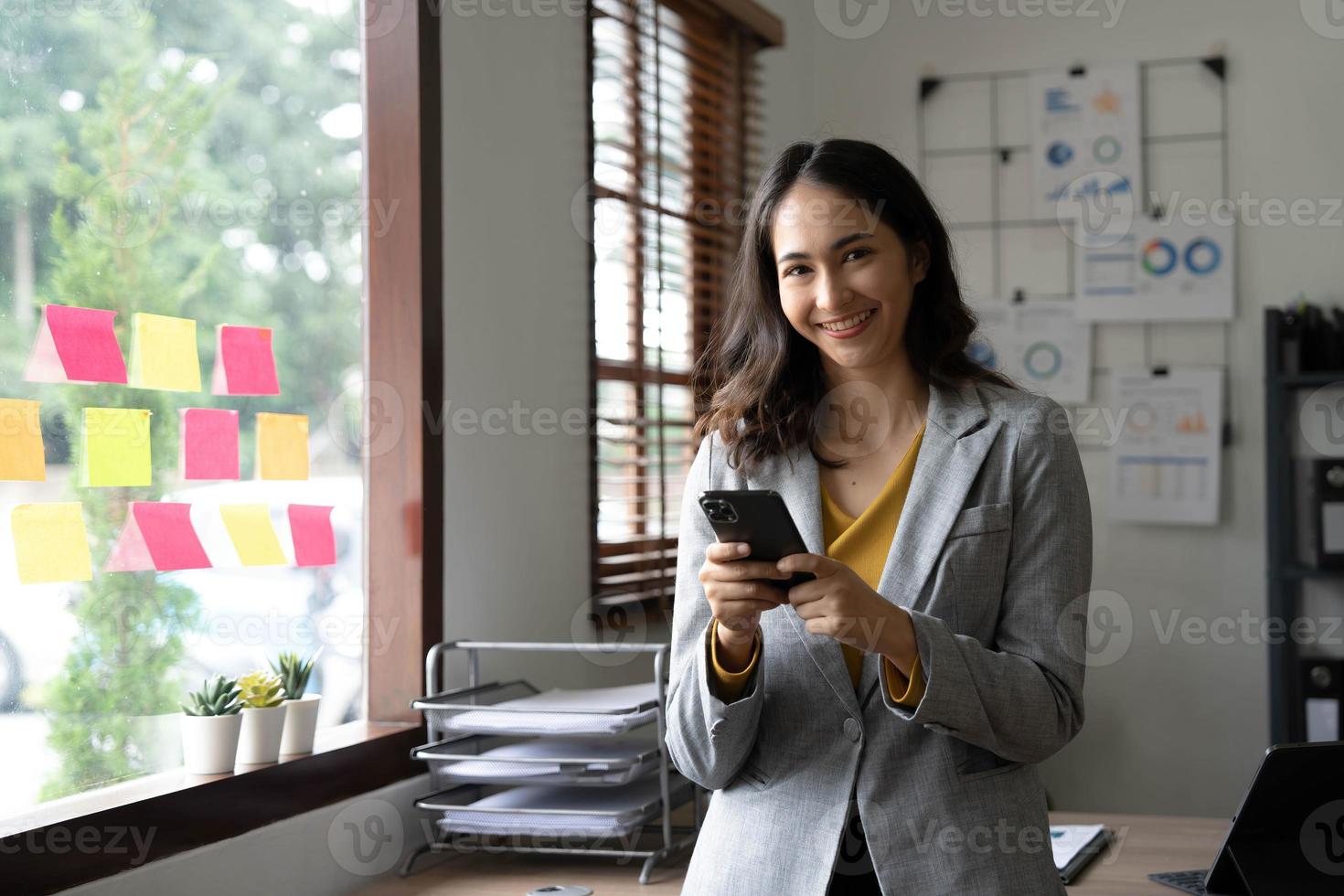 sorridente bellissimo asiatico donna d'affari analizzando grafico e grafico mostrando i cambiamenti su il mercato e Tenere smartphone a ufficio. foto