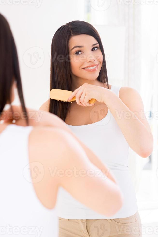 donna pettinatura capelli. attraente giovane donna pettinatura sua capelli mentre guardare a il specchio foto