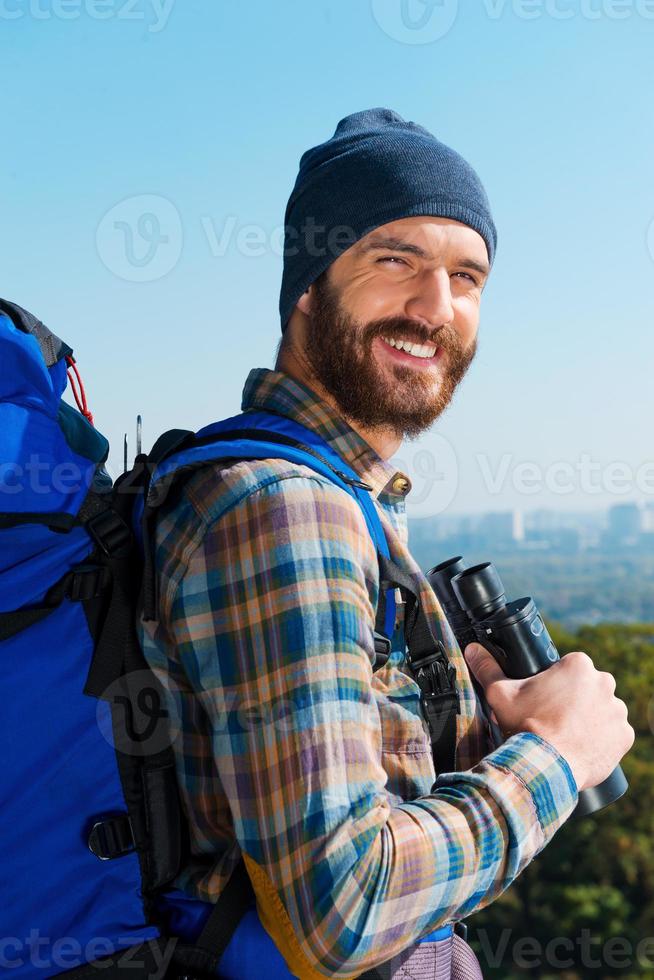 su il superiore. bello giovane uomo trasporto zaino e guardare a telecamera attraverso il le spalle con Sorridi mentre in piedi nel il natura e Tenere binocolo foto