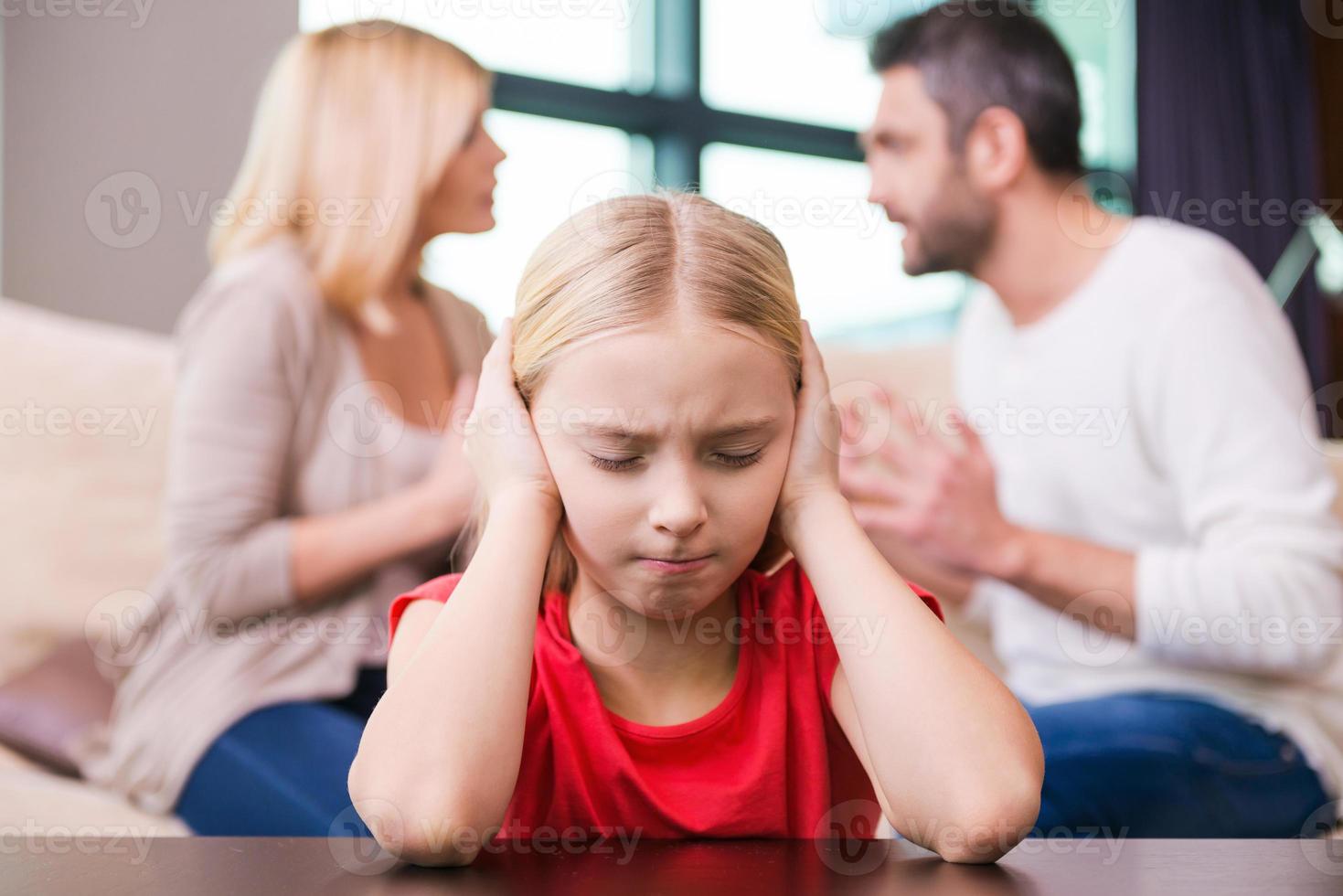 per favore fermare combattente depresso poco ragazza pendente a il tavolo e copertura orecchio con mani mentre sua genitori urlando a ogni altro nel il sfondo foto