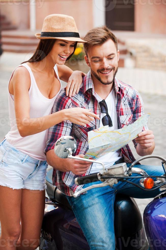 io pensare noi siamo Qui bellissimo giovane amorevole coppia seduta su scooter insieme e l'esame carta geografica mentre donna puntamento esso e sorridente foto