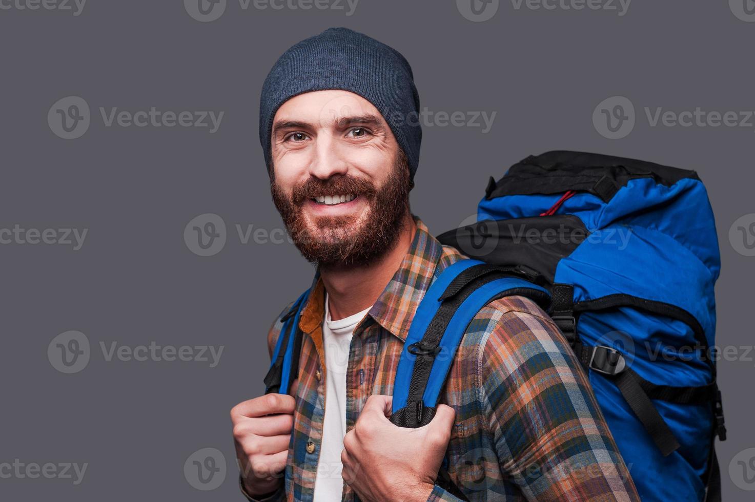 contento turista. bello giovane barbuto uomo trasporto zaino e sorridente mentre in piedi contro grigio sfondo foto