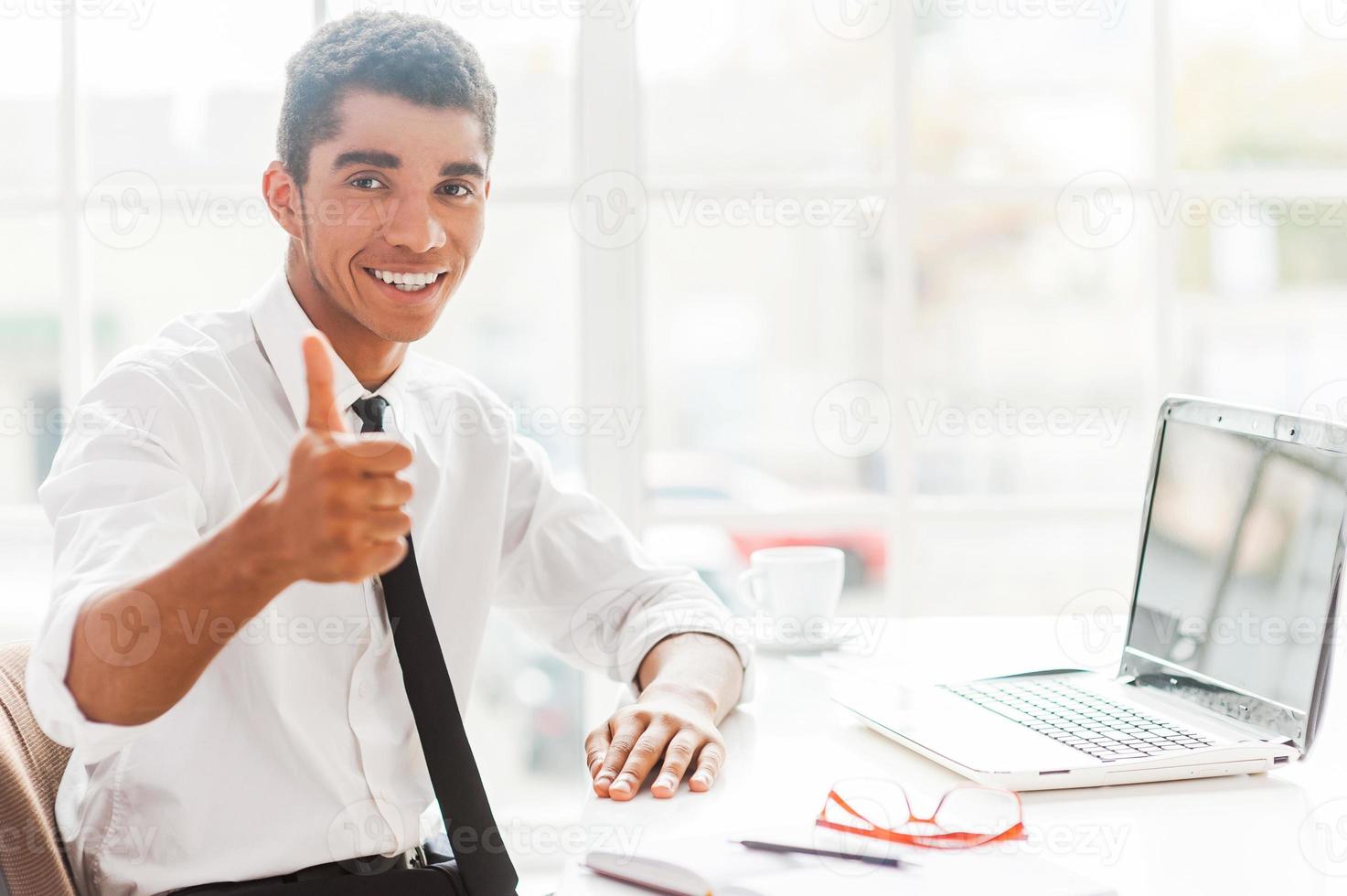 bene lavoro fiducioso giovane afro americano uomo nel vestito formale allungamento su mano con pollice su mentre seduta a il suo opera posto foto