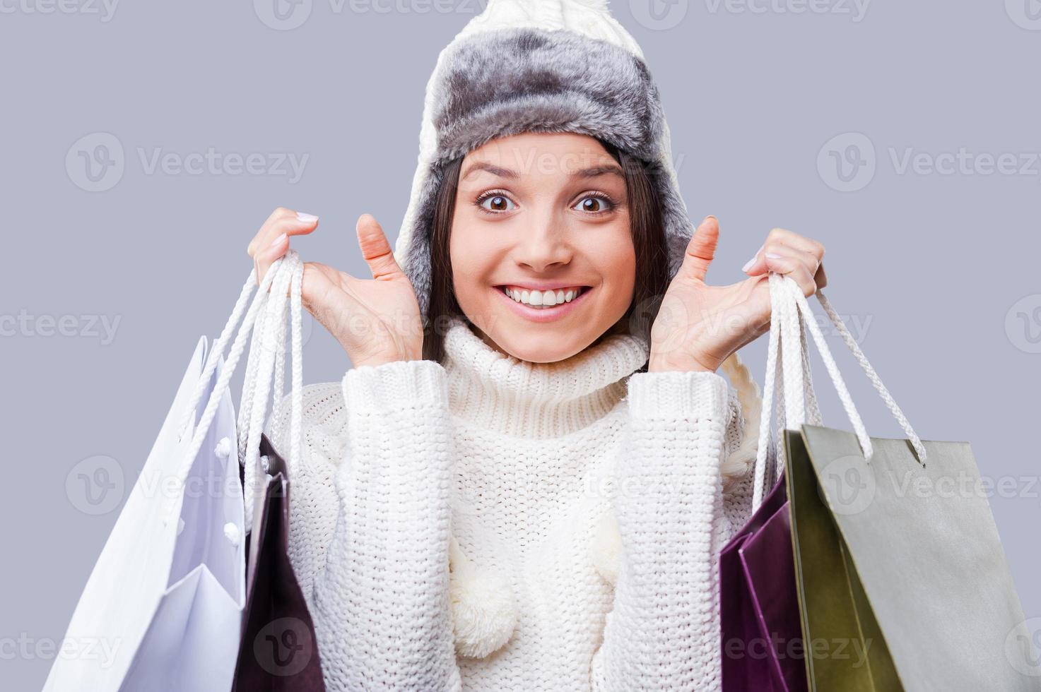 shopping nel qualunque tempo metereologico . contento giovane donne indossare caldo inverno capi di abbigliamento e Tenere pacchi con acquisti mentre in piedi contro grigio sfondo foto