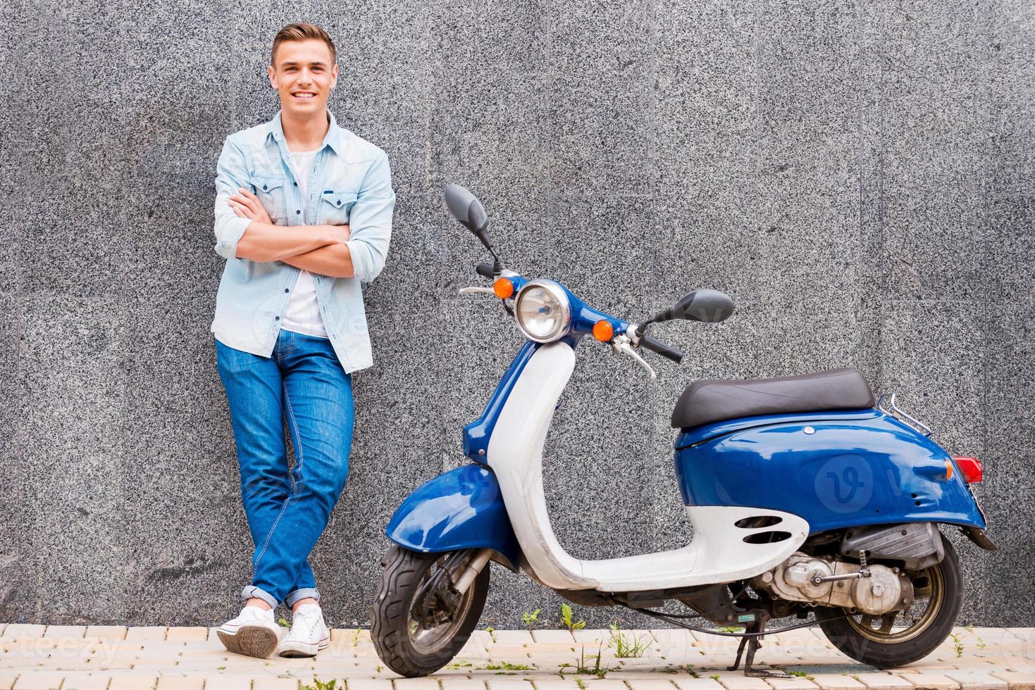 me e mio nuovo scooter. bello giovane uomo pendente a il parete e sorridente mentre in piedi vicino il suo nuovo scooter foto