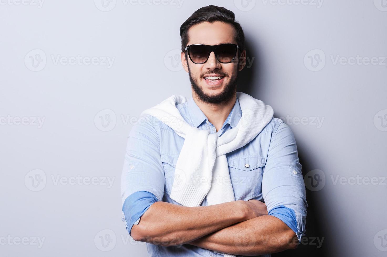 affascinante bello. bello giovane uomo nel blu camicia conservazione braccia attraversato e sorridente a telecamera mentre in piedi contro grigio sfondo foto