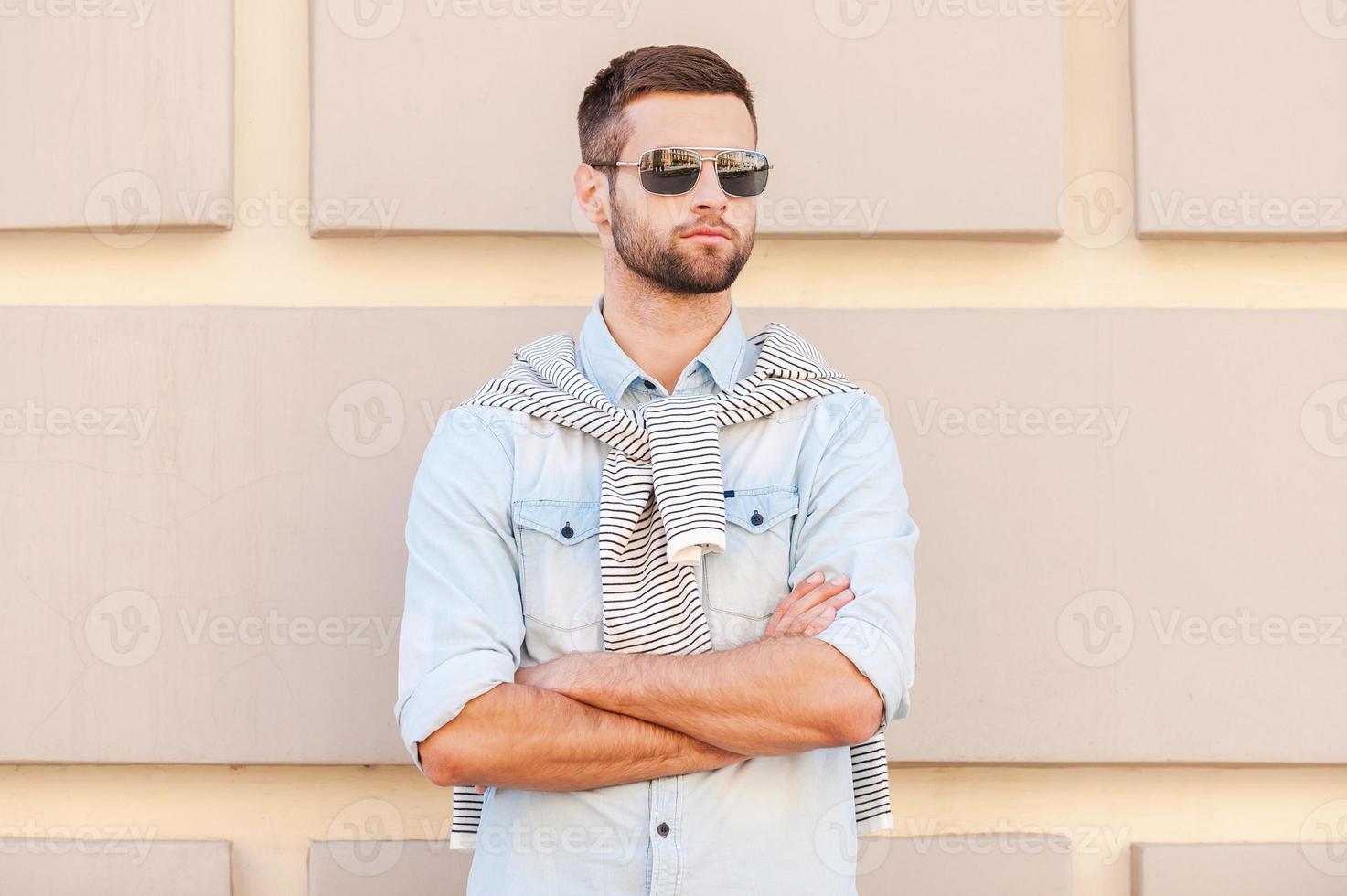 freddo e bello. bello giovane uomo nel occhiali da sole conservazione braccia attraversato e guardare lontano mentre in piedi nel davanti di il strutturato parete all'aperto foto