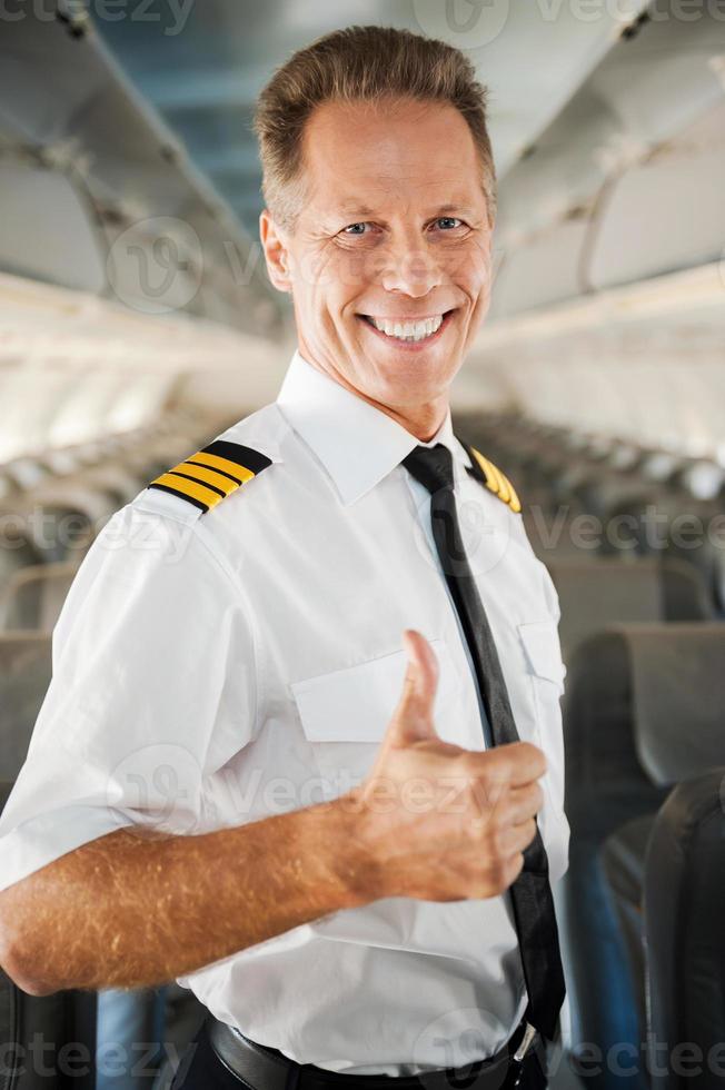io amore mio lavoro fiducioso maschio pilota nel uniforme mostrando il suo pollice su e sorridente mentre in piedi dentro di il aereo foto