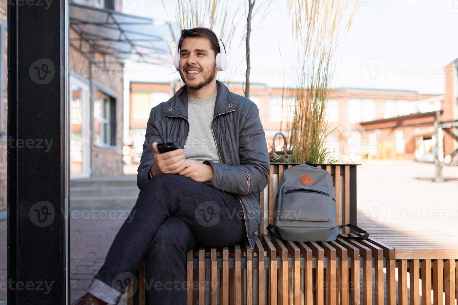 adulto uomo ascoltando per musica su cuffie con un' Telefono nel il suo mani seduta su un' panchina Il prossimo per un ufficio edificio foto