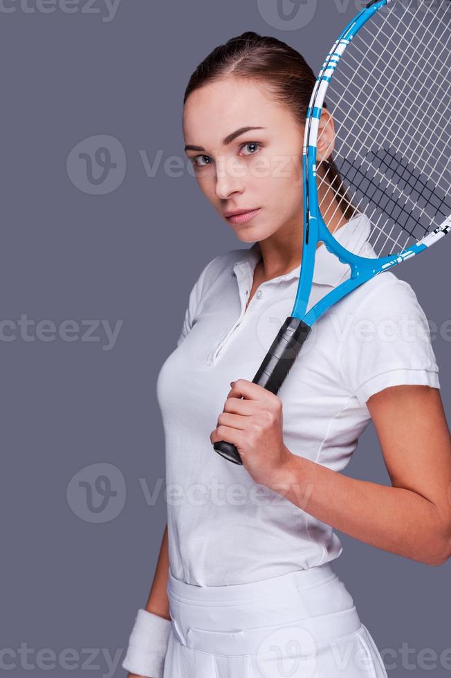 grave competitivo. bellissimo giovane donne nel gli sport Abiti Tenere tennis racchetta su sua spalla e guardare a telecamera mentre in piedi contro grigio sfondo foto