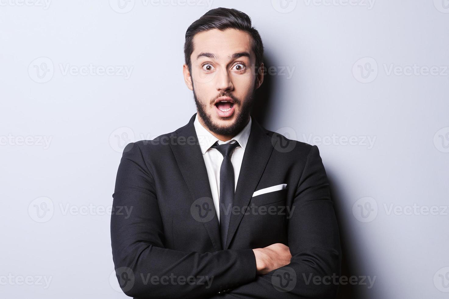 quello è incredibile sorpreso giovane uomo nel vestito formale conservazione bocca Aperto e guardare a telecamera mentre in piedi contro grigio sfondo foto