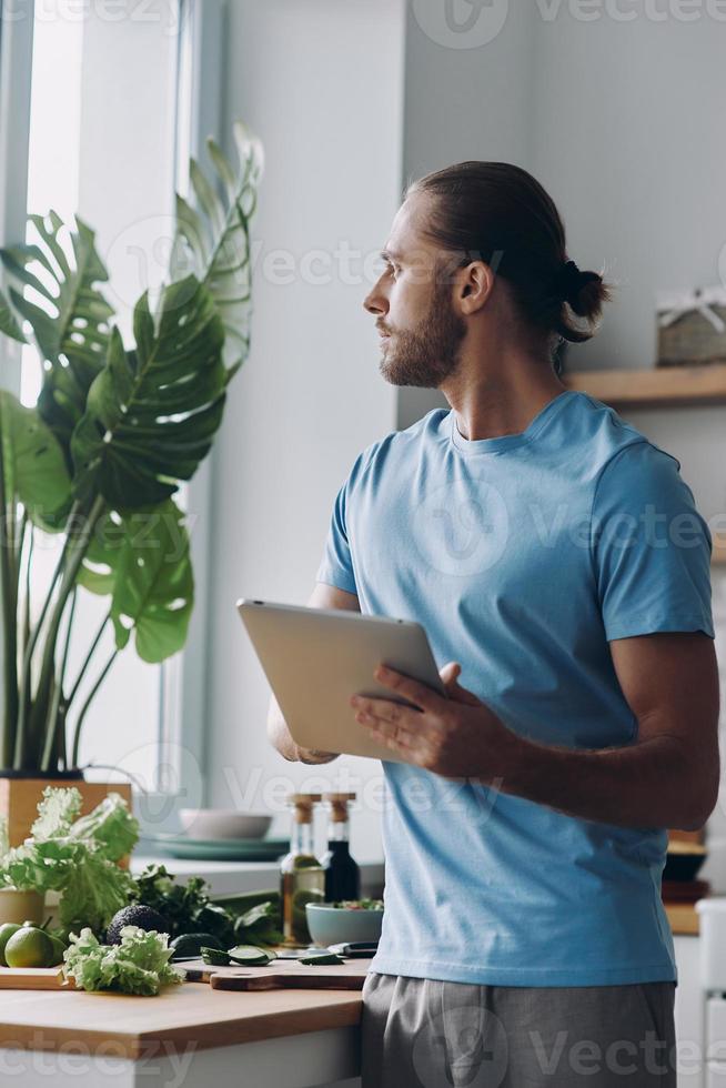 riflessivo giovane uomo utilizzando digitale tavoletta mentre preparazione cibo a il domestico cucina foto