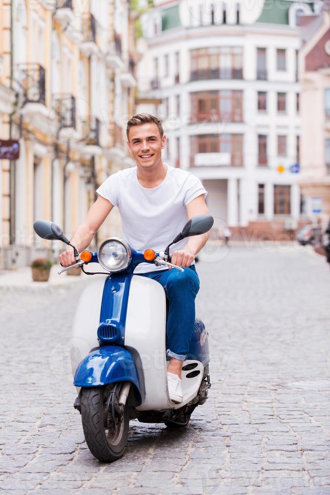 appena equitazione intorno a. bello giovane uomo nel occhiali da sole equitazione scooter lungo il strada e sorridente foto