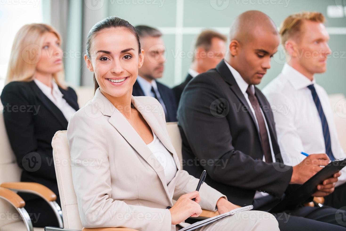 donna a il conferenza. gruppo di attività commerciale persone nel vestito formale seduta a il sedie nel conferenza sala e scrittura qualcosa Gin loro Nota pastiglie mentre attraente donna guardare a telecamera e sorridente foto