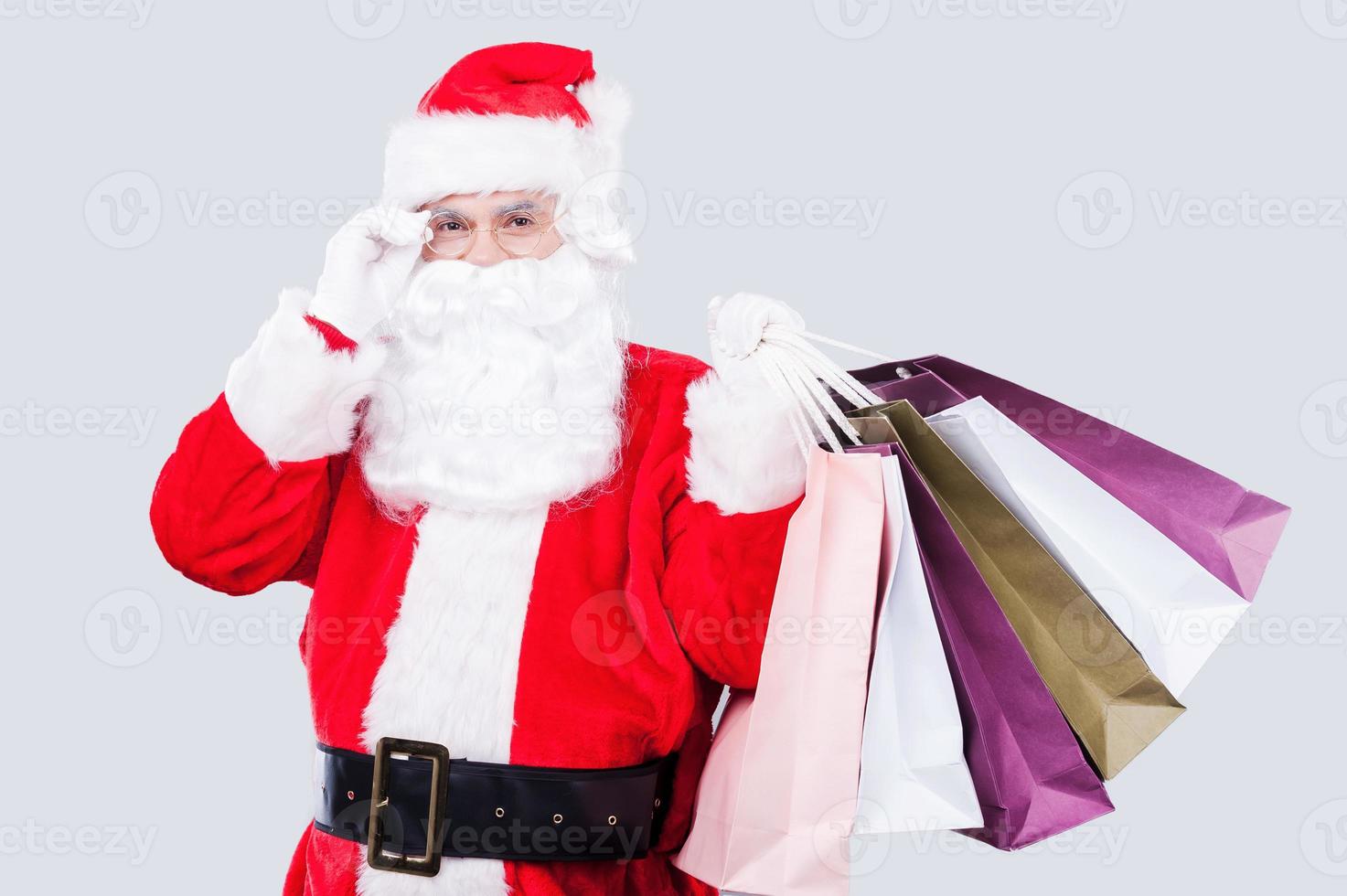 pronto per Natale i saldi contento Santa Claus Tenere shopping borse e regolazione il suo occhiali mentre in piedi contro grigio sfondo foto