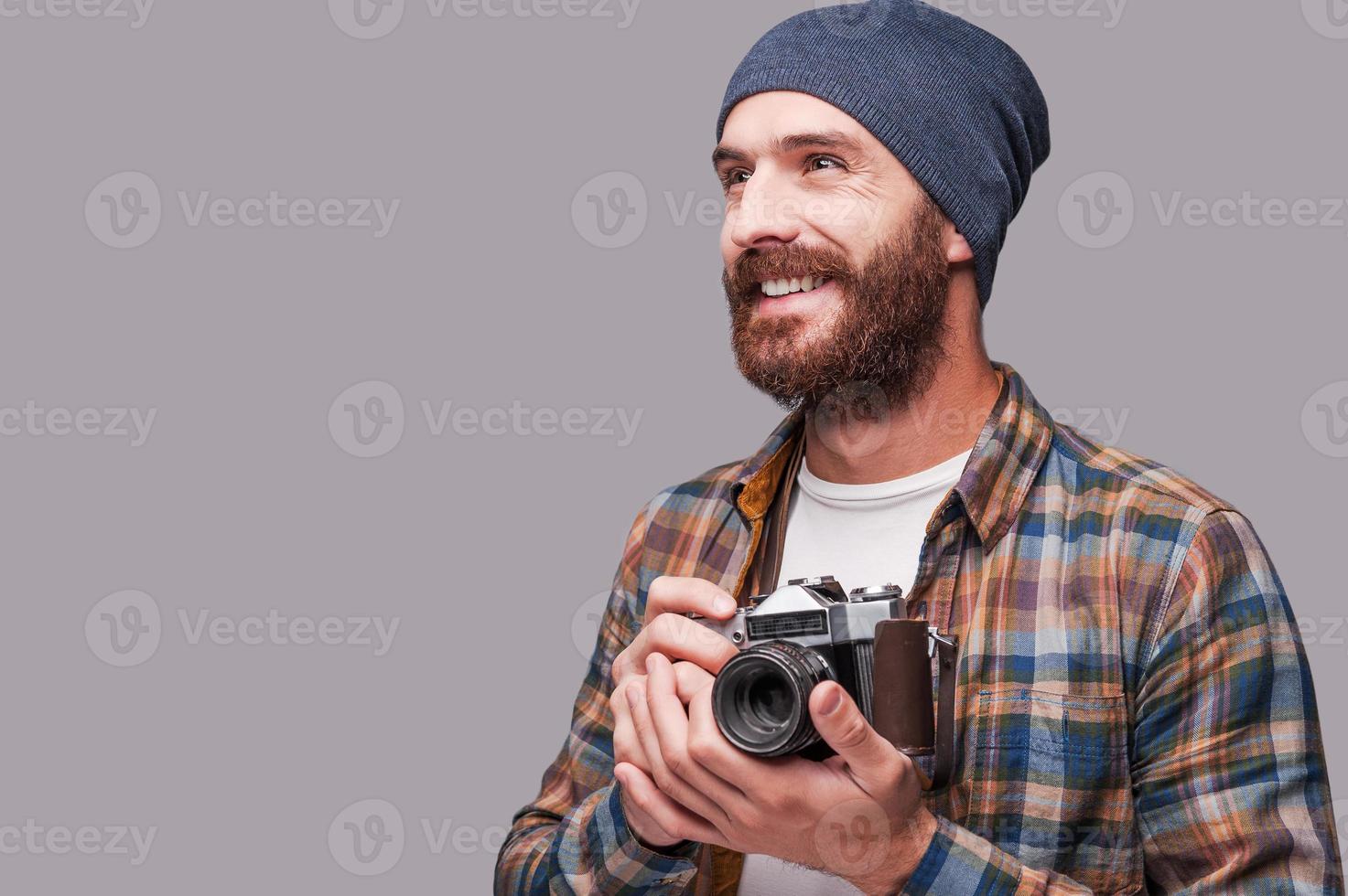 contento fotografo. bello giovane barbuto uomo Tenere vecchio stile telecamera e guardare lontano con Sorridi mentre in piedi contro grigio sfondo foto