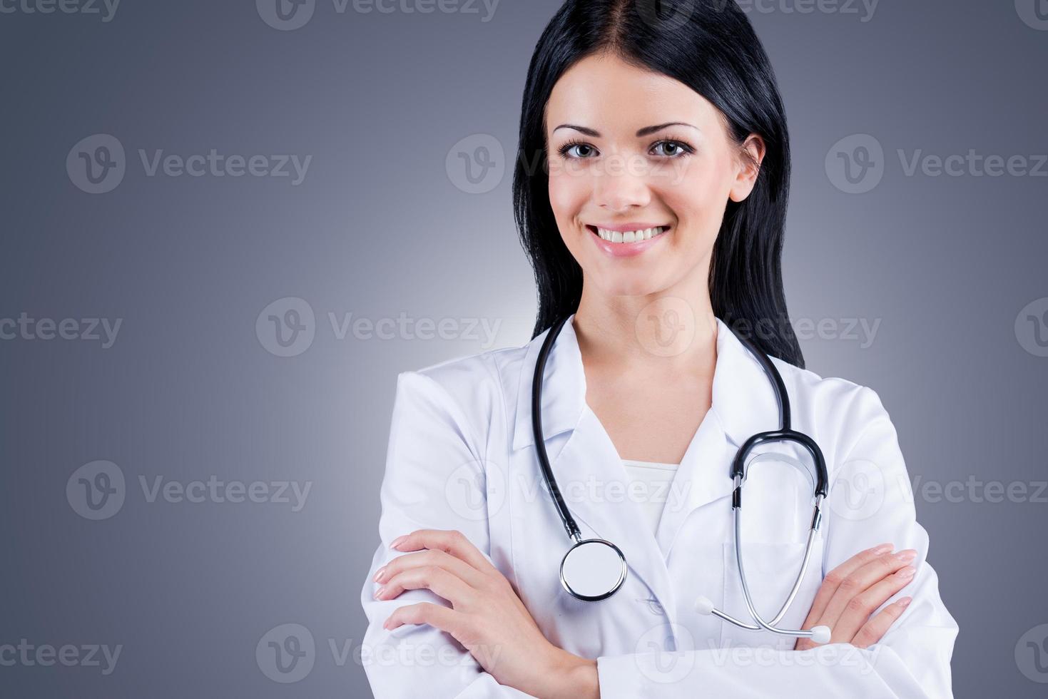 il tuo Salute è nel il bene mani. fiducioso femmina medico nel bianca uniforme guardare a telecamera e sorridente mentre in piedi contro grigio sfondo foto