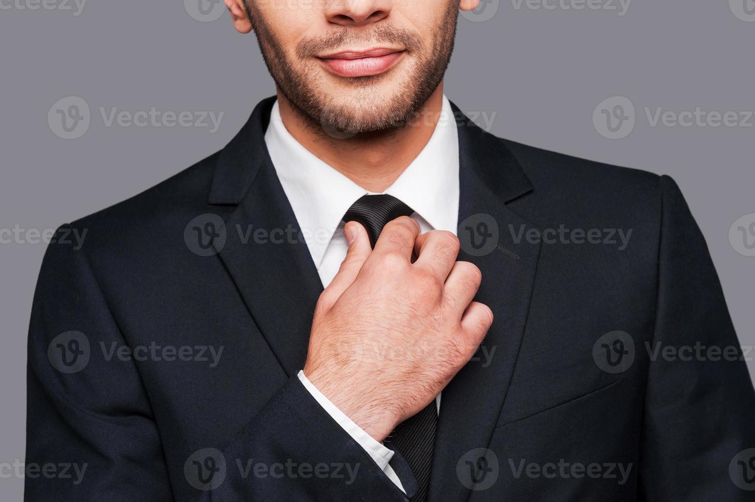 bianca collare fiducia. avvicinamento di giovane uomo nel vestito formale regolazione il suo cravatta mentre in piedi contro grigio sfondo foto