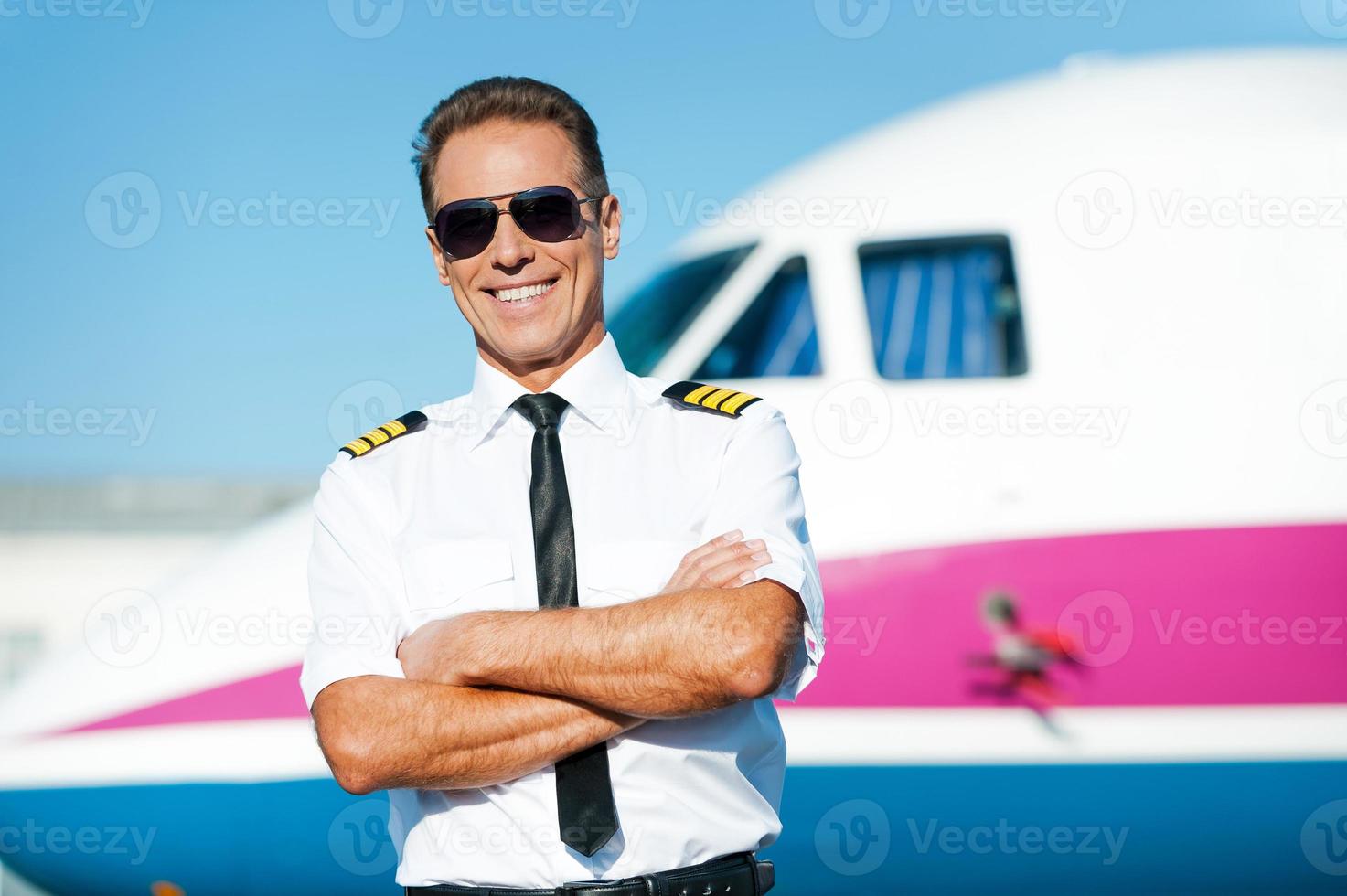 fiducioso pilota. fiducioso maschio pilota nel uniforme conservazione braccia attraversato e sorridente con aereo nel il sfondo foto