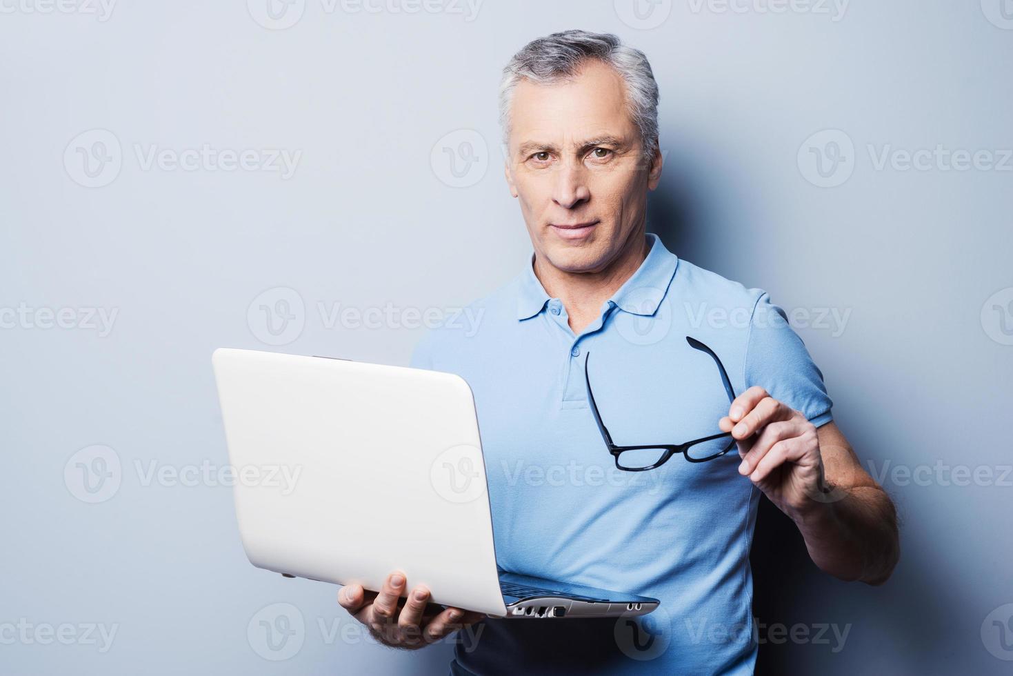 sempre pronto per Aiuto voi. allegro anziano uomo nel casuale Tenere il suo occhiali e il computer portatile mentre in piedi contro grigio sfondo foto