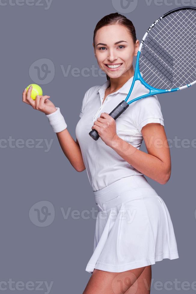 portando il bellezza per il gioco. bellissimo giovane donne nel gli sport Abiti Tenere tennis racchetta su sua spalla e sorridente mentre in piedi contro grigio sfondo foto