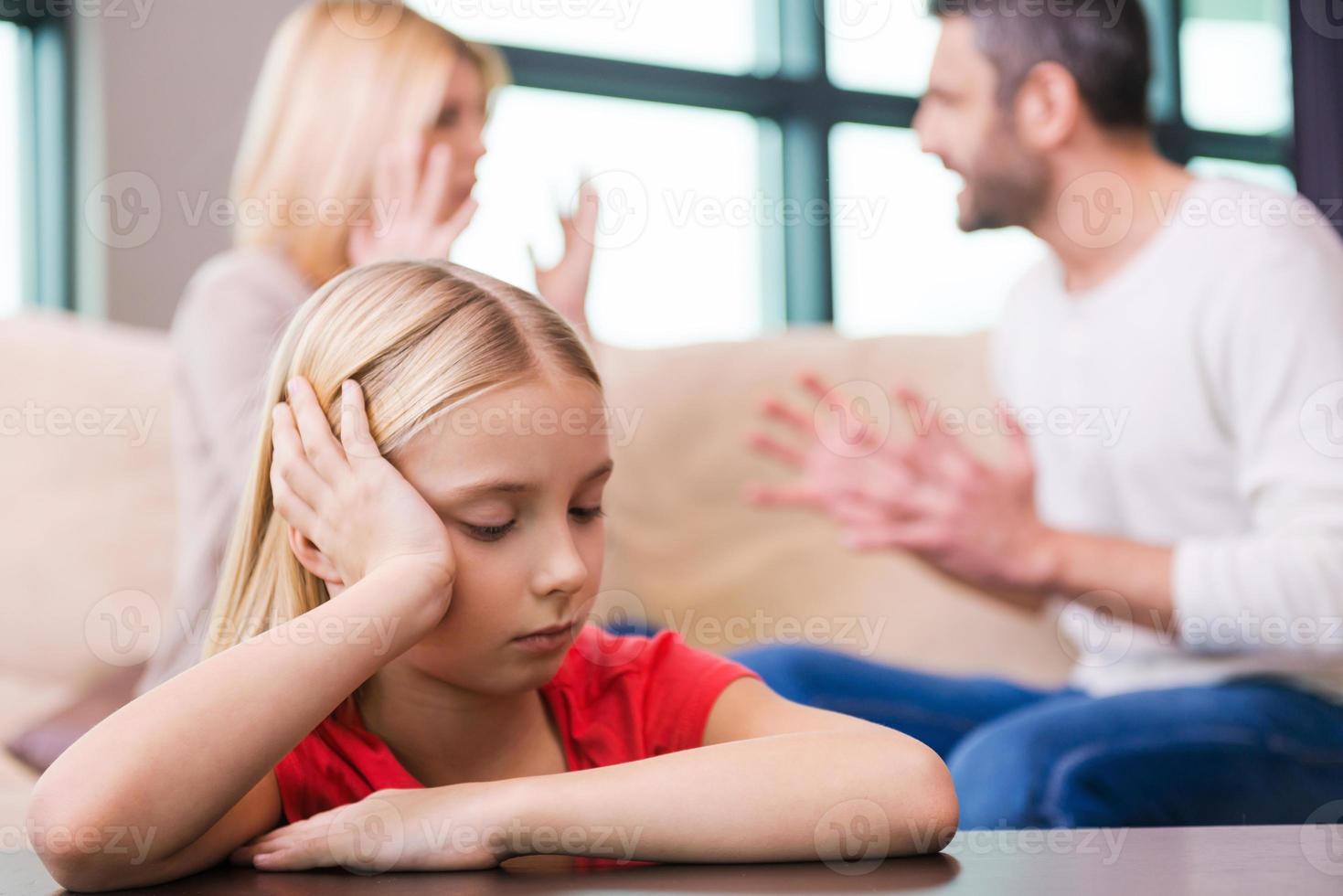 io speranza essi volontà opera esso fuori. triste poco ragazza pendente a il tavolo e Tenere testa nel mano mentre sua genitori urlando a ogni altro nel il sfondo foto