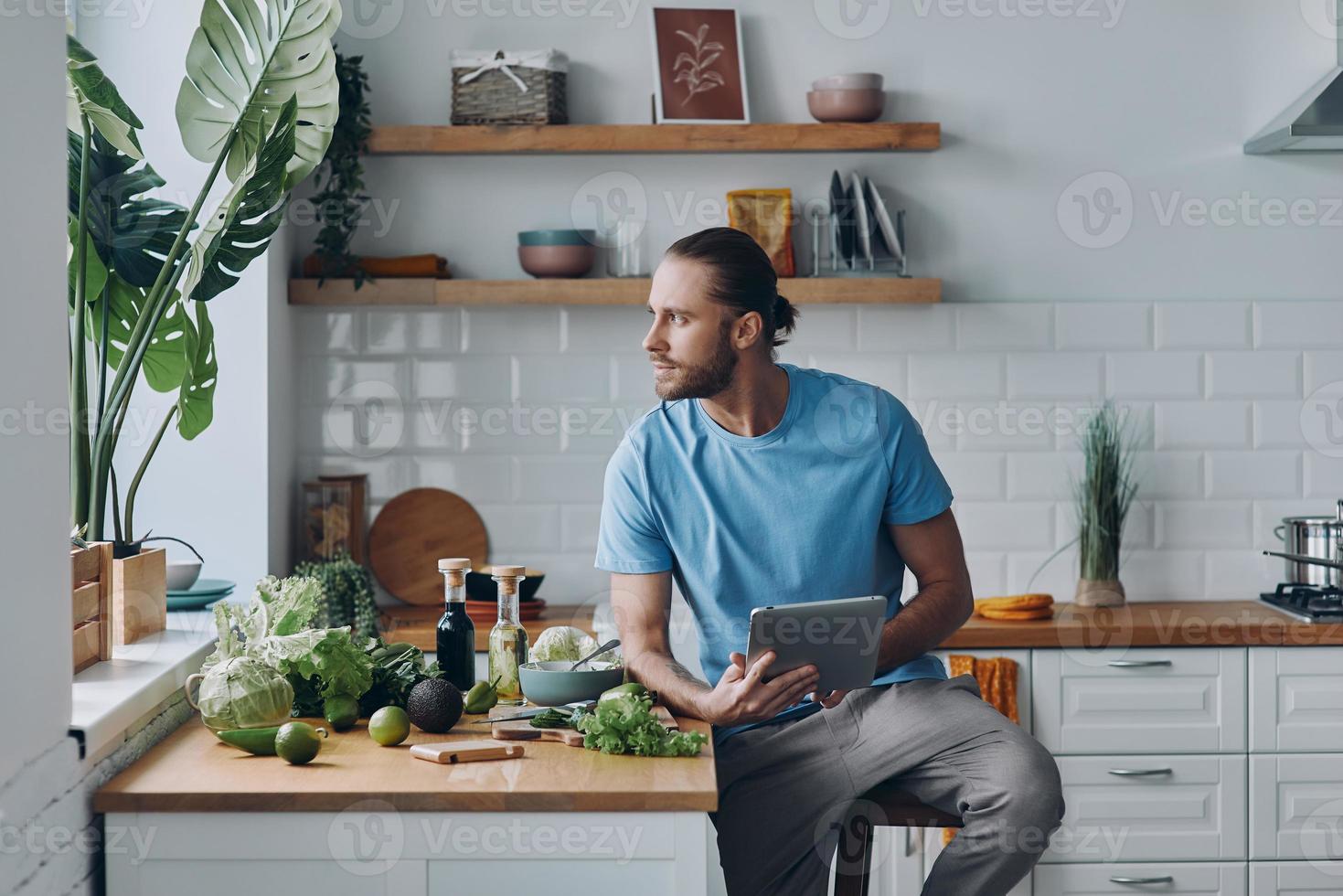 bello giovane uomo Tenere digitale tavoletta mentre preparazione cibo a il domestico cucina foto