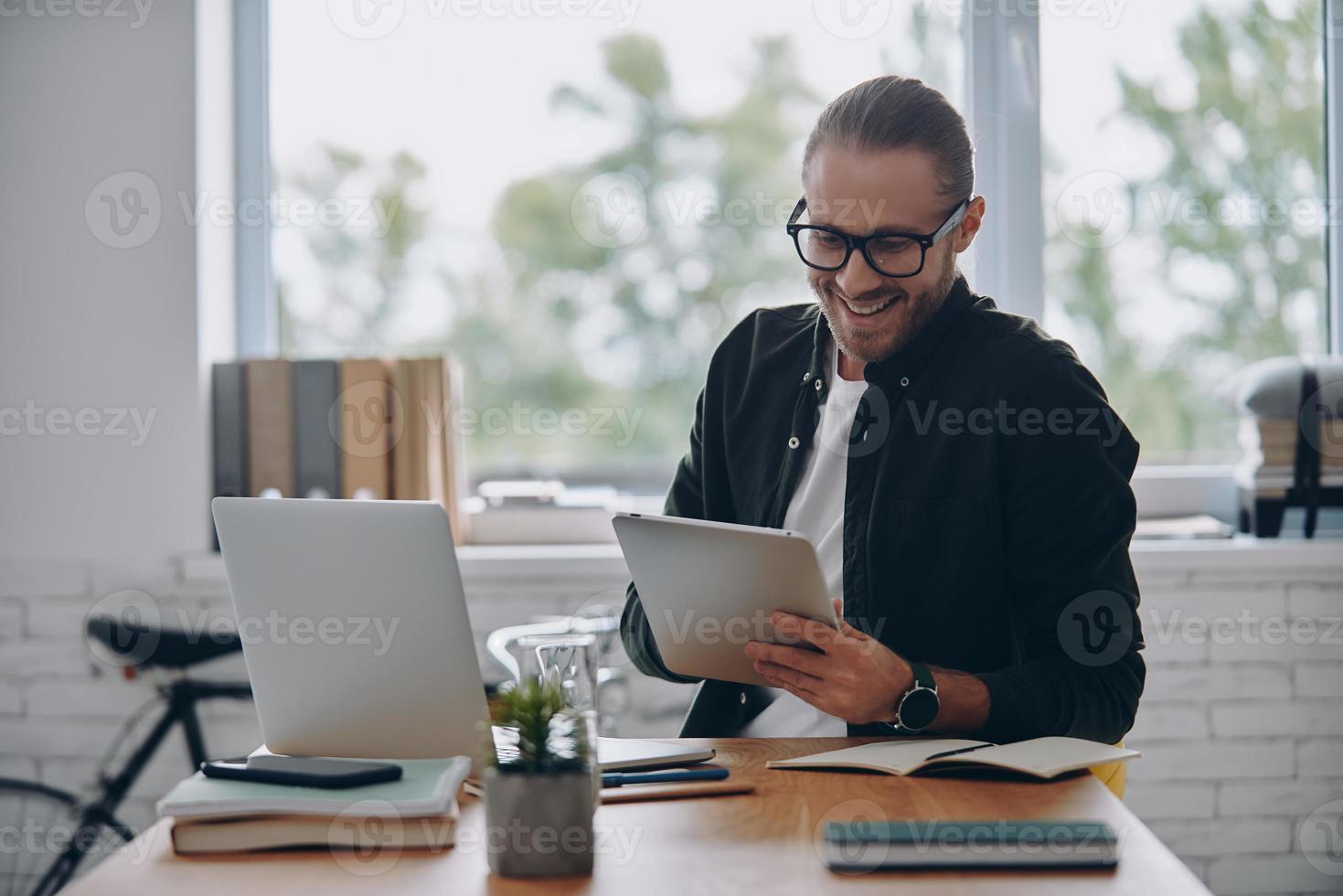 allegro giovane uomo utilizzando digitale tavoletta mentre seduta a il suo Lavorando posto nel ufficio foto