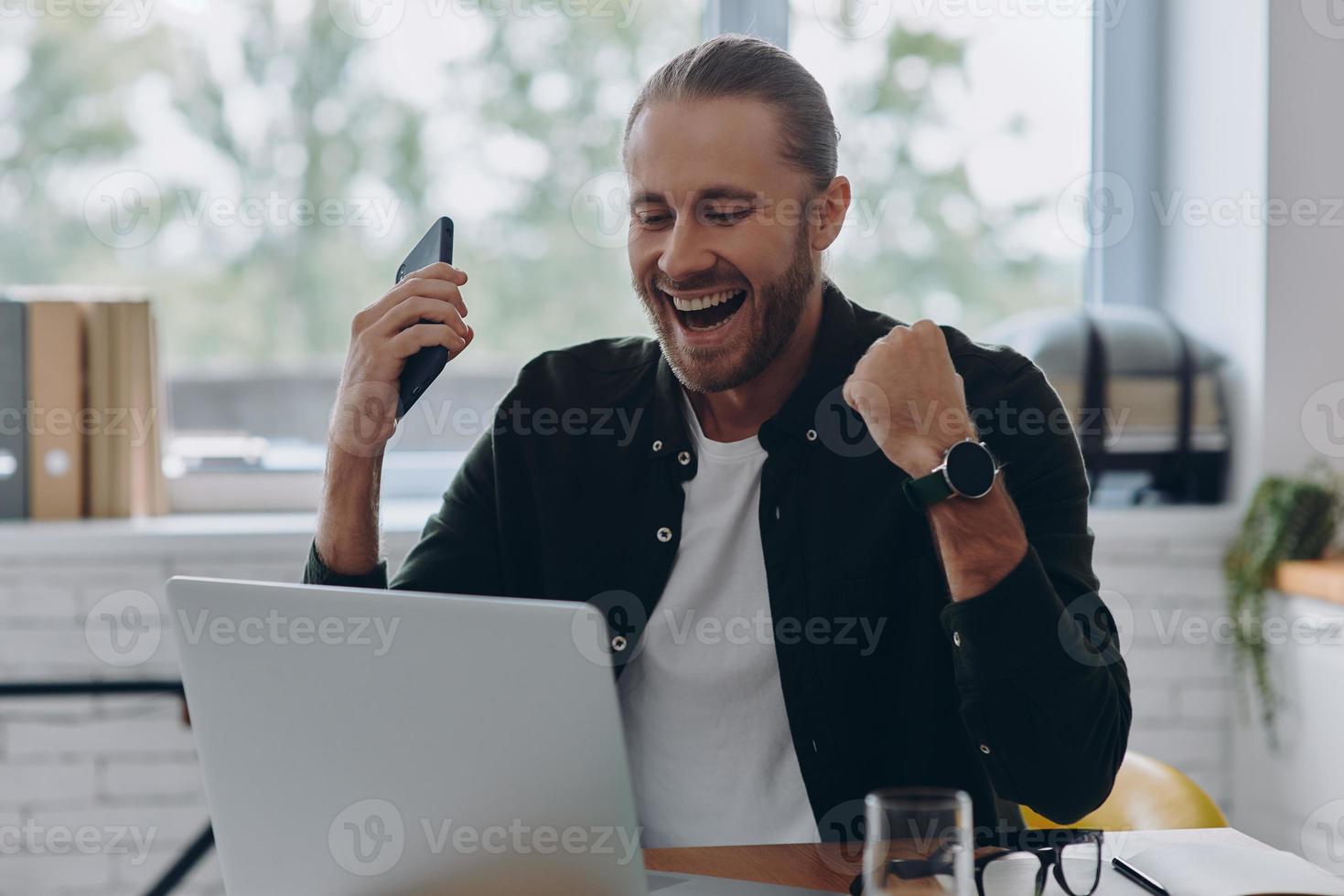 contento giovane uomo guardare a il computer portatile e Esprimere a gesti mentre seduta a il suo Lavorando posto nel ufficio foto