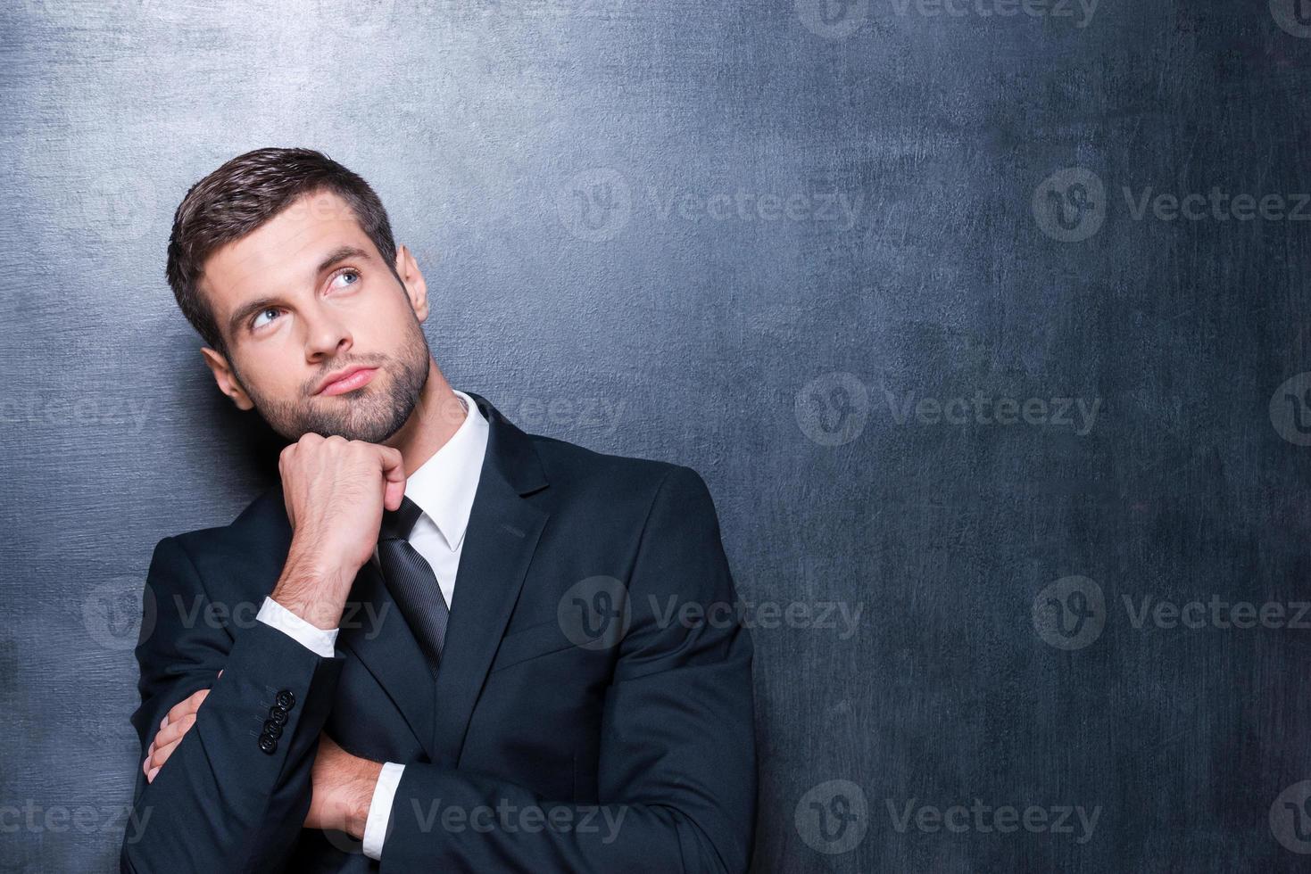 riflessivo uomo d'affari. bello giovane uomo nel camicia e cravatta guardare lontano e Tenere mano su mento mentre in piedi contro lavagna foto