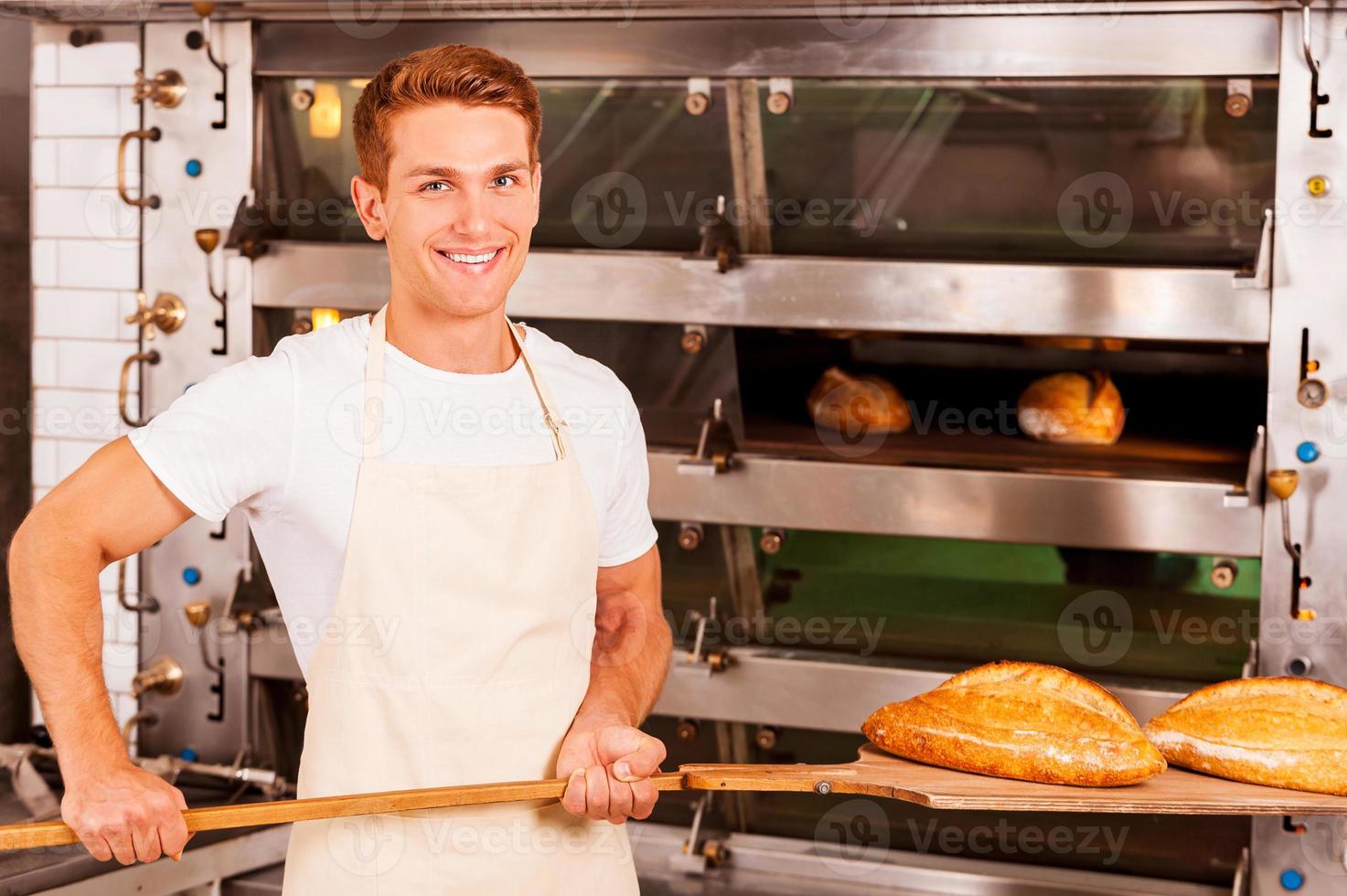 fresco al forno pane per voi. fiducioso giovane uomo nel grembiule assunzione fresco al forno pane a partire dal forno e sorridente foto