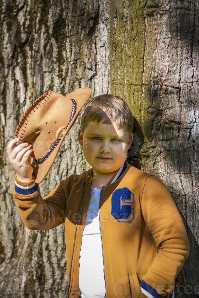 carino ragazzo in posa nel un' cowboy cappello nel il boschi di un' albero. il di sole raggi avvolgere il spazio. interazione storia per il libro. spazio per copiatura foto